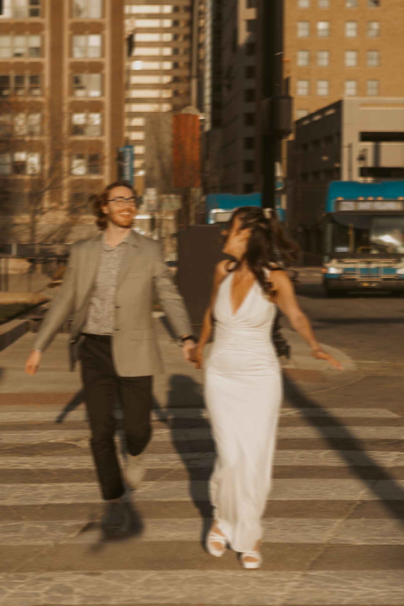 Joyful couple holding hands and running across a city crosswalk, woman in white dress and man in gray blazer.