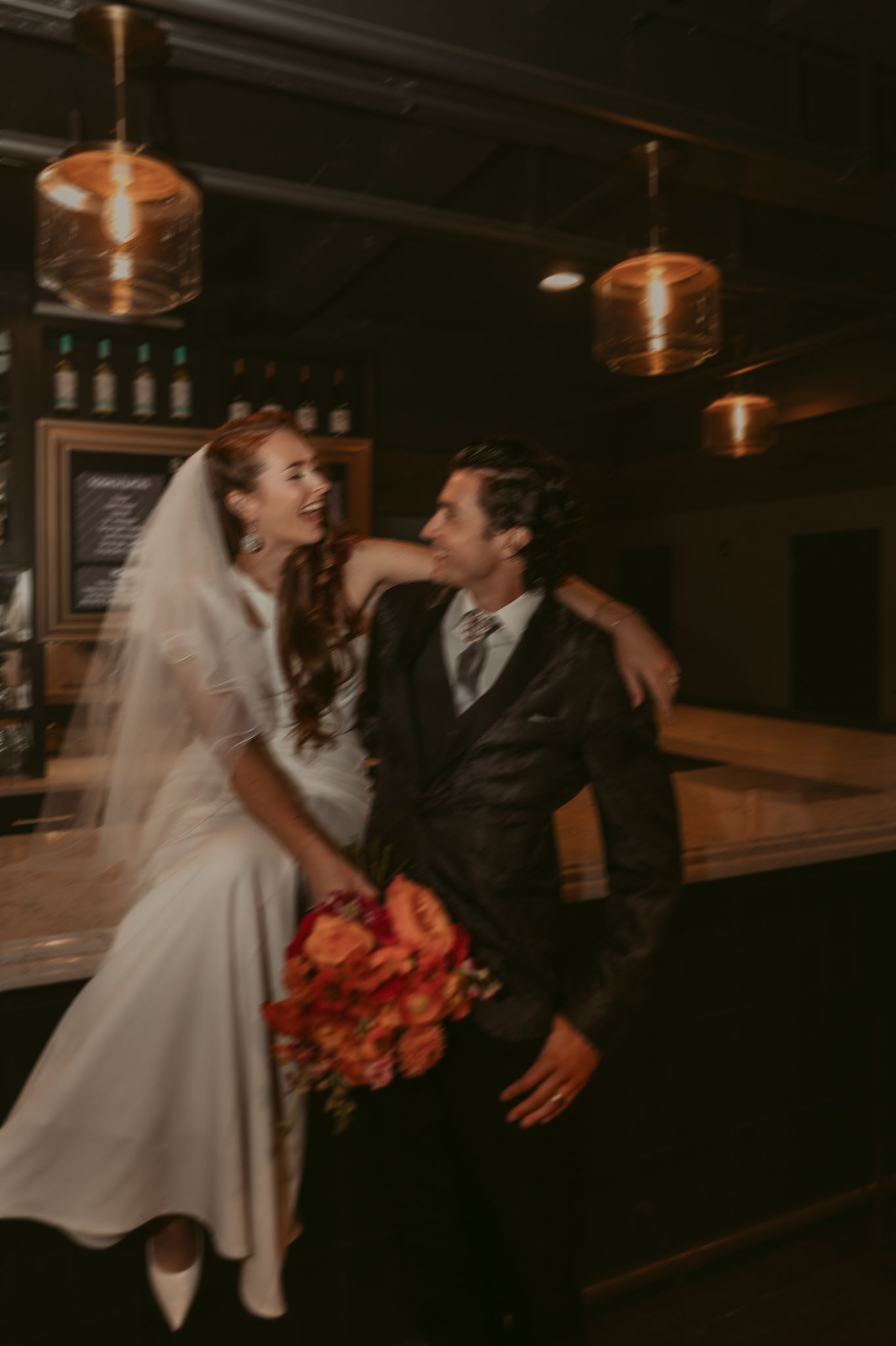 Laughing bride in veil and groom in dark suit embrace at a warmly lit bar with pendant lights.