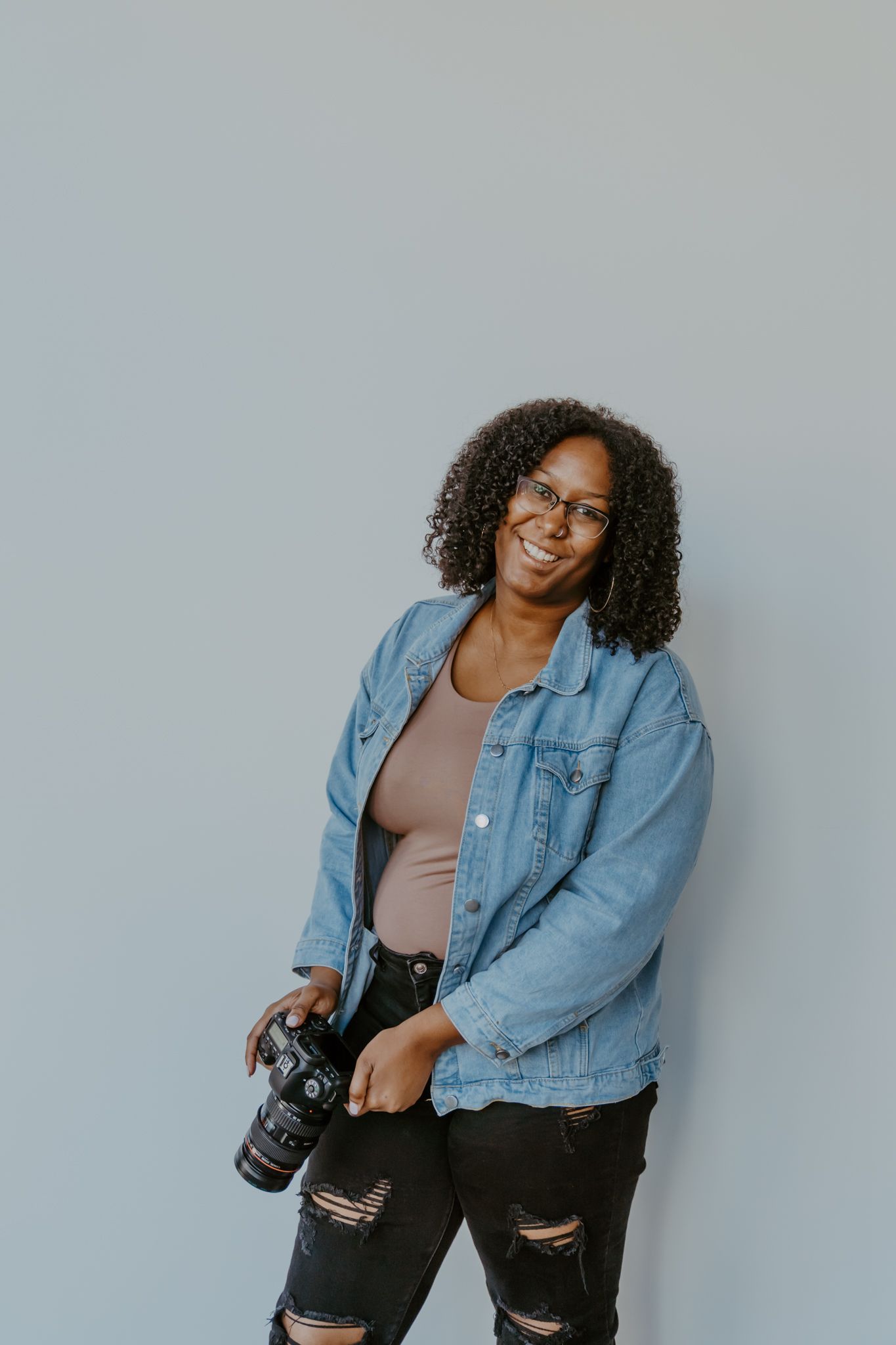 Smiling female photographer in denim jacket holding a DSLR camera against a light gray wall.