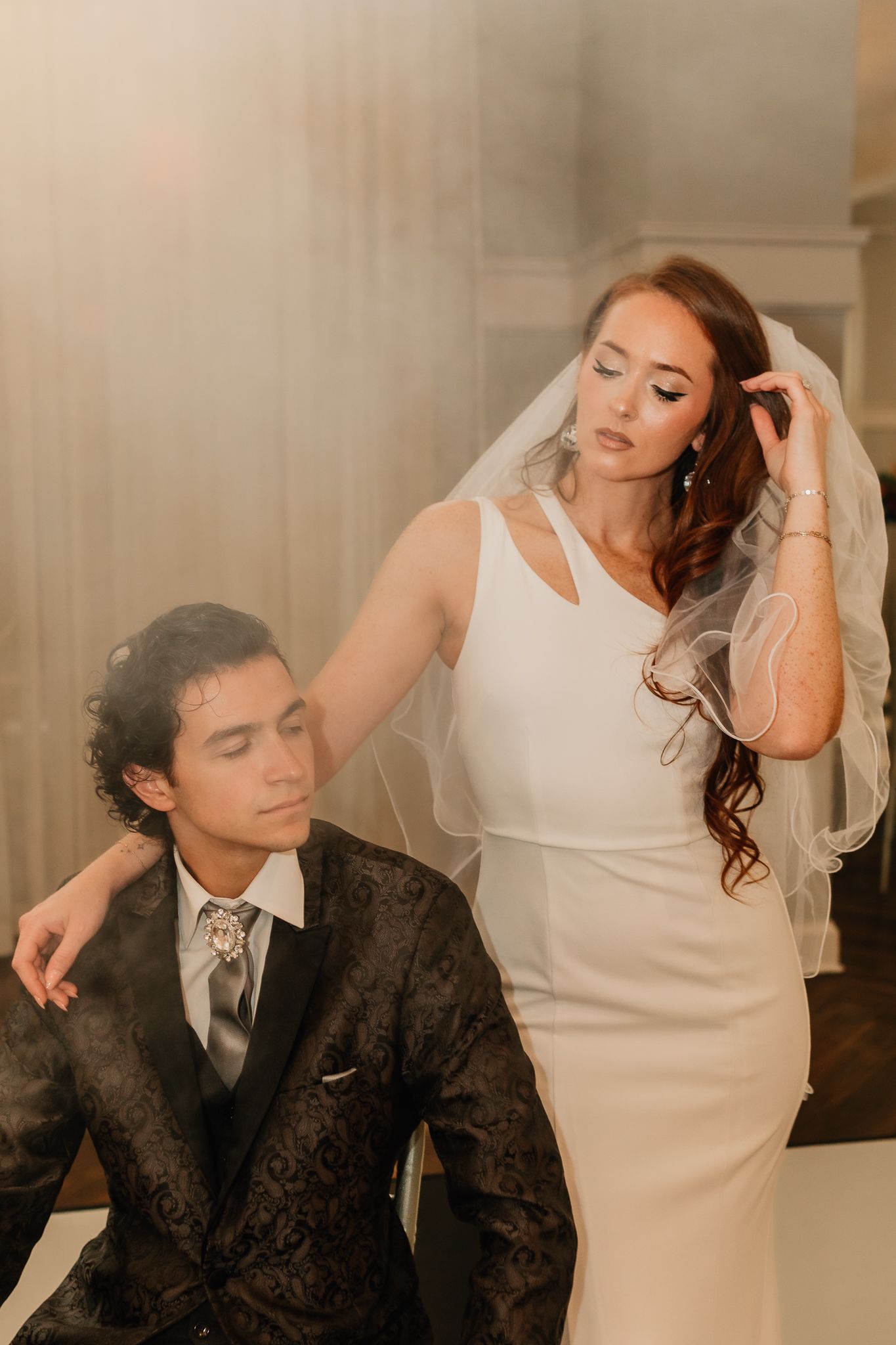 Elegant bride in sleek white gown with veil rests hand on seated groom in ornate dark suit indoors.