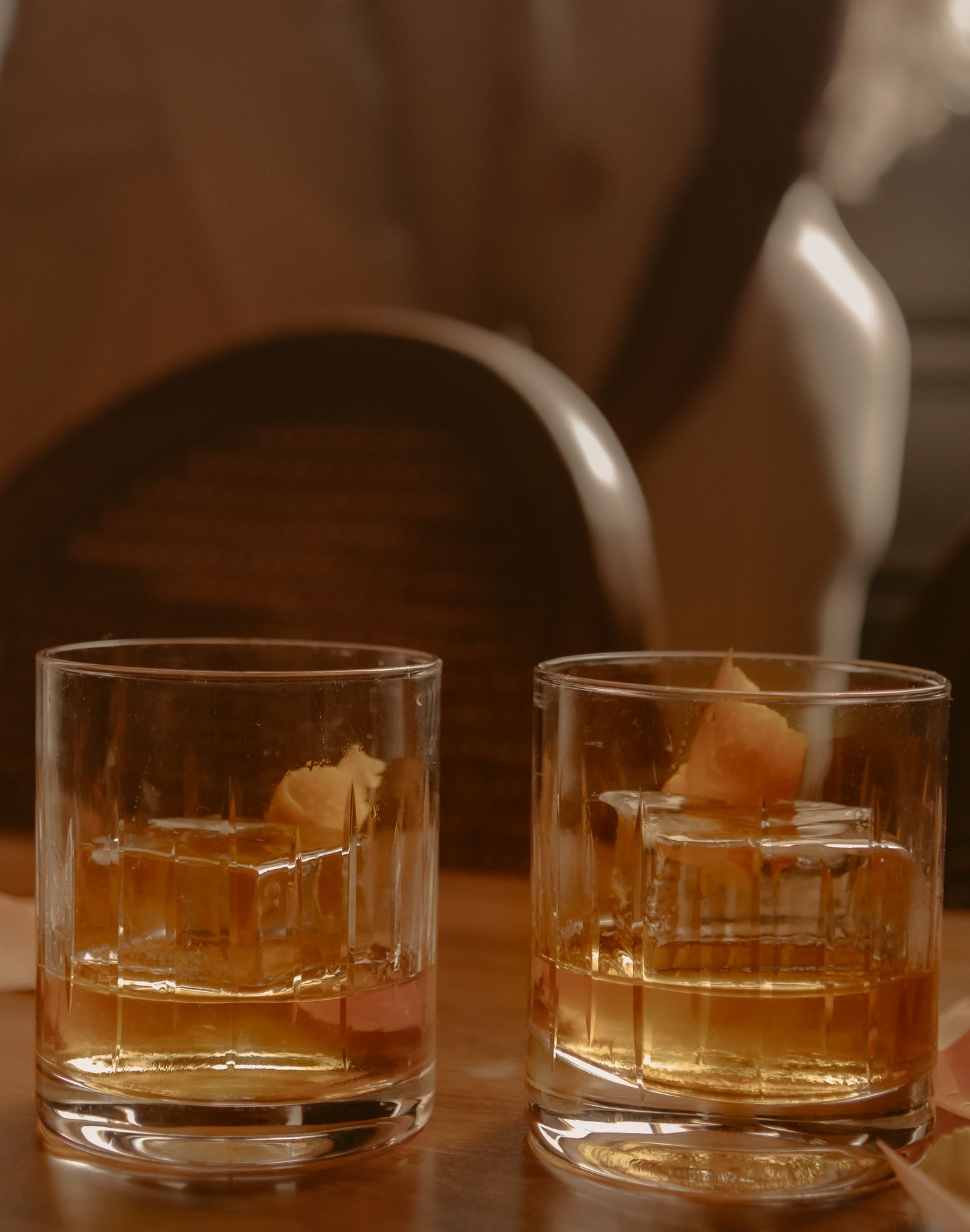 Two whiskey old fashioned cocktails with large ice cubes on wooden table in warm light.