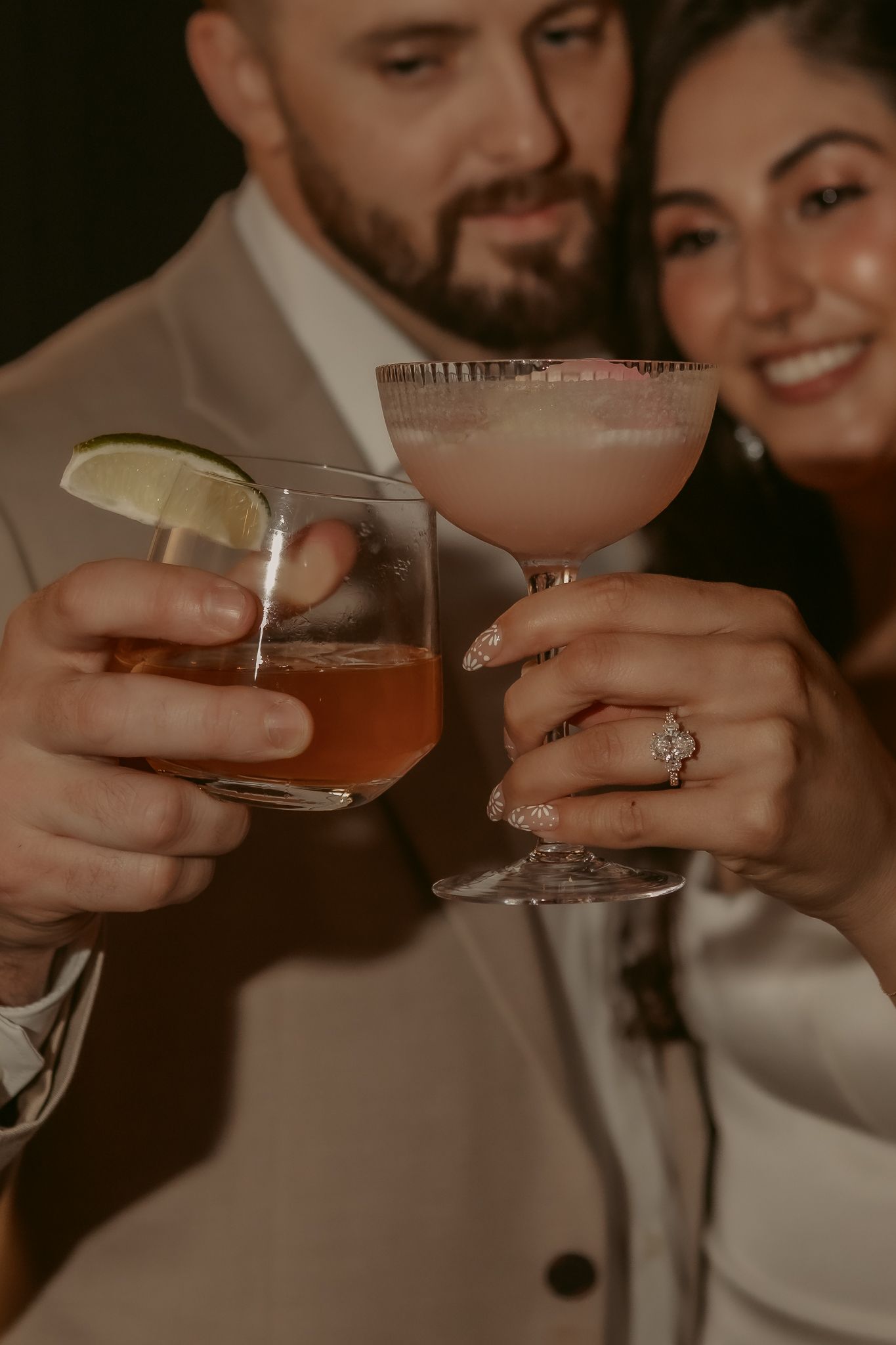 Wedding couple toasting with cocktails, bride wearing diamond engagement ring in romantic moody lighting.