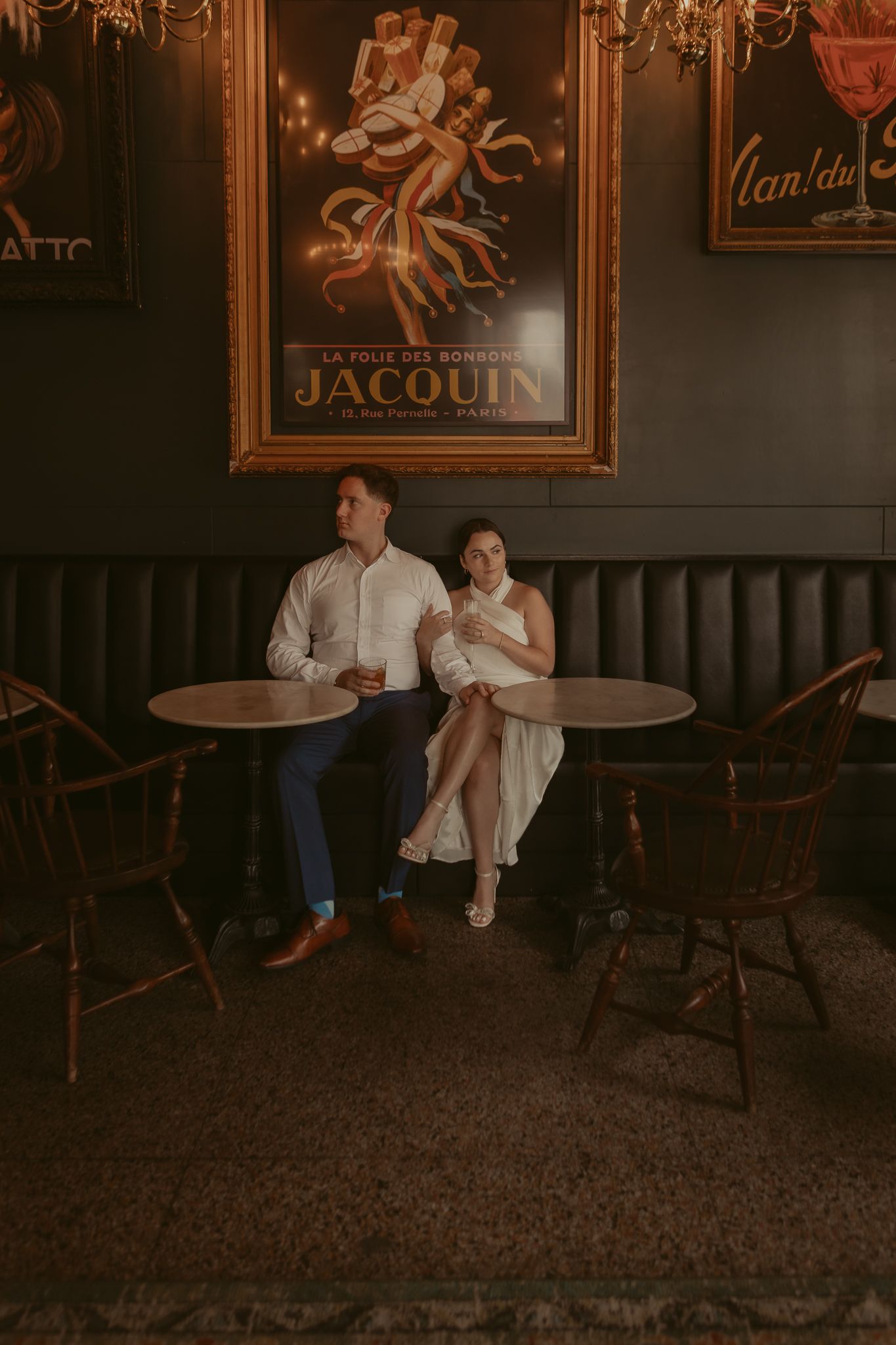 Bride and groom sit together in dark vintage bar beneath ornate Jacquin Paris posters.