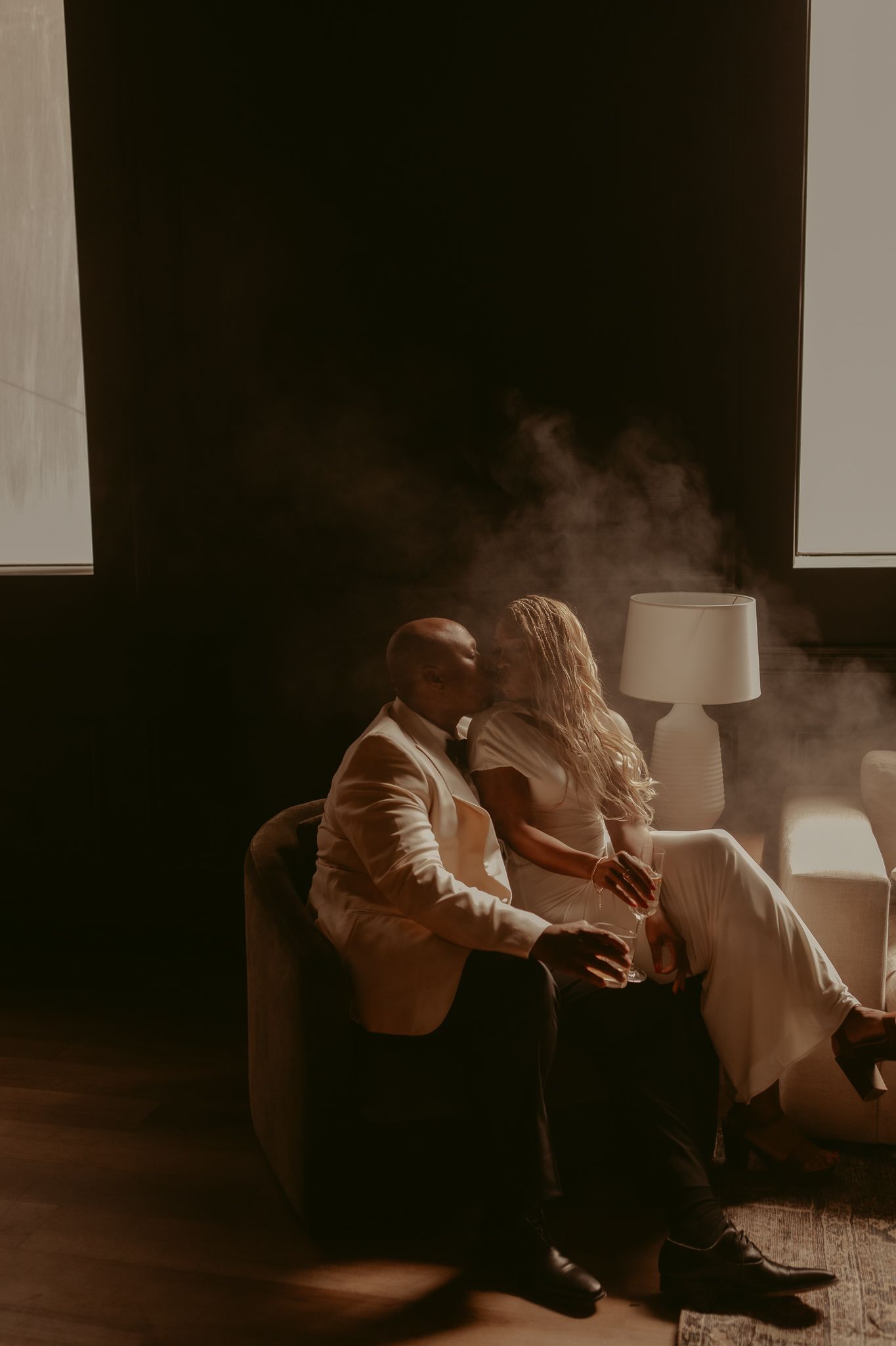 Newlywed couple sitting intimately in armchair with dramatic moody lighting and smoke.