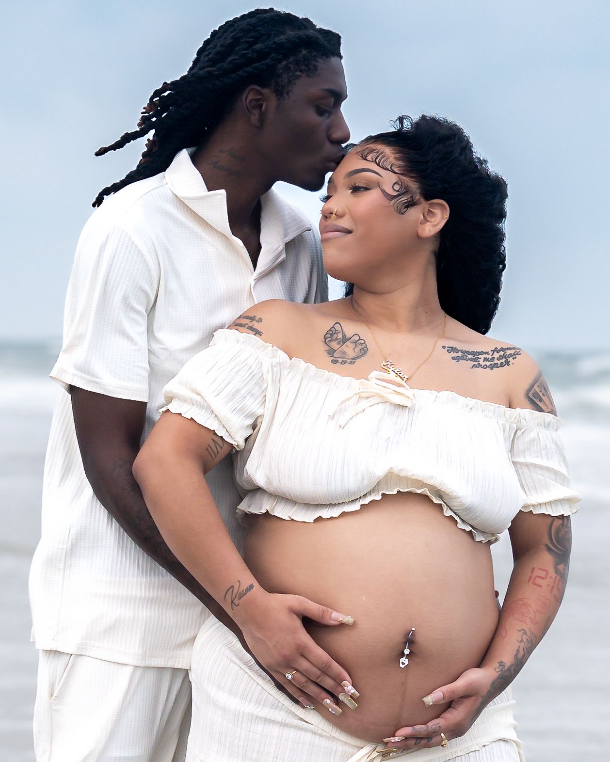 Couple in white outfits sharing a tender maternity beach photo, woman with tattoos cradling baby bump.
