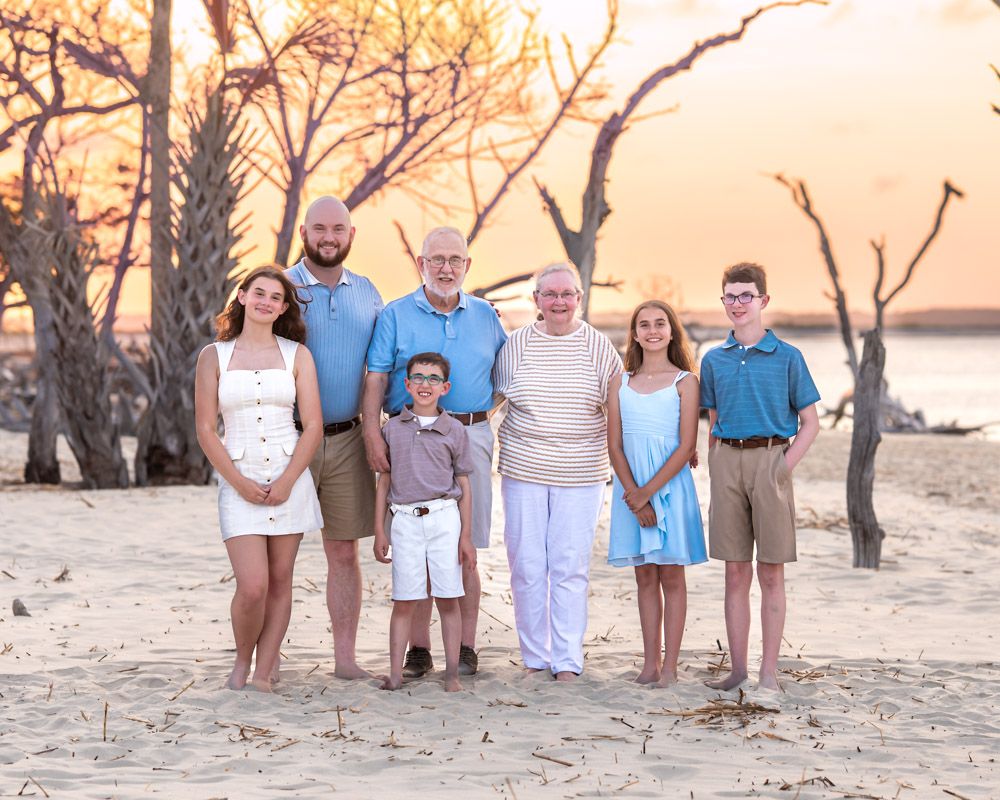 Multi-generational family posing together on a sandy beach at sunset with bare trees in the background.