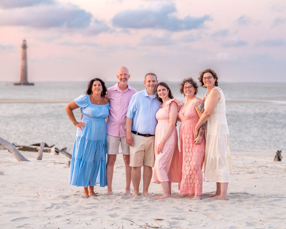 Six people pose together on a sandy beach at sunset with a lighthouse in the background.