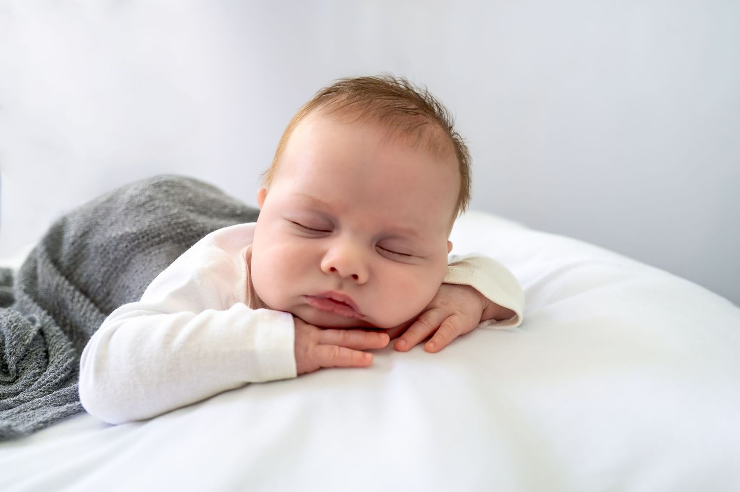 Peaceful newborn baby sleeping on white pillow with hands under chin, wrapped in gray blanket.