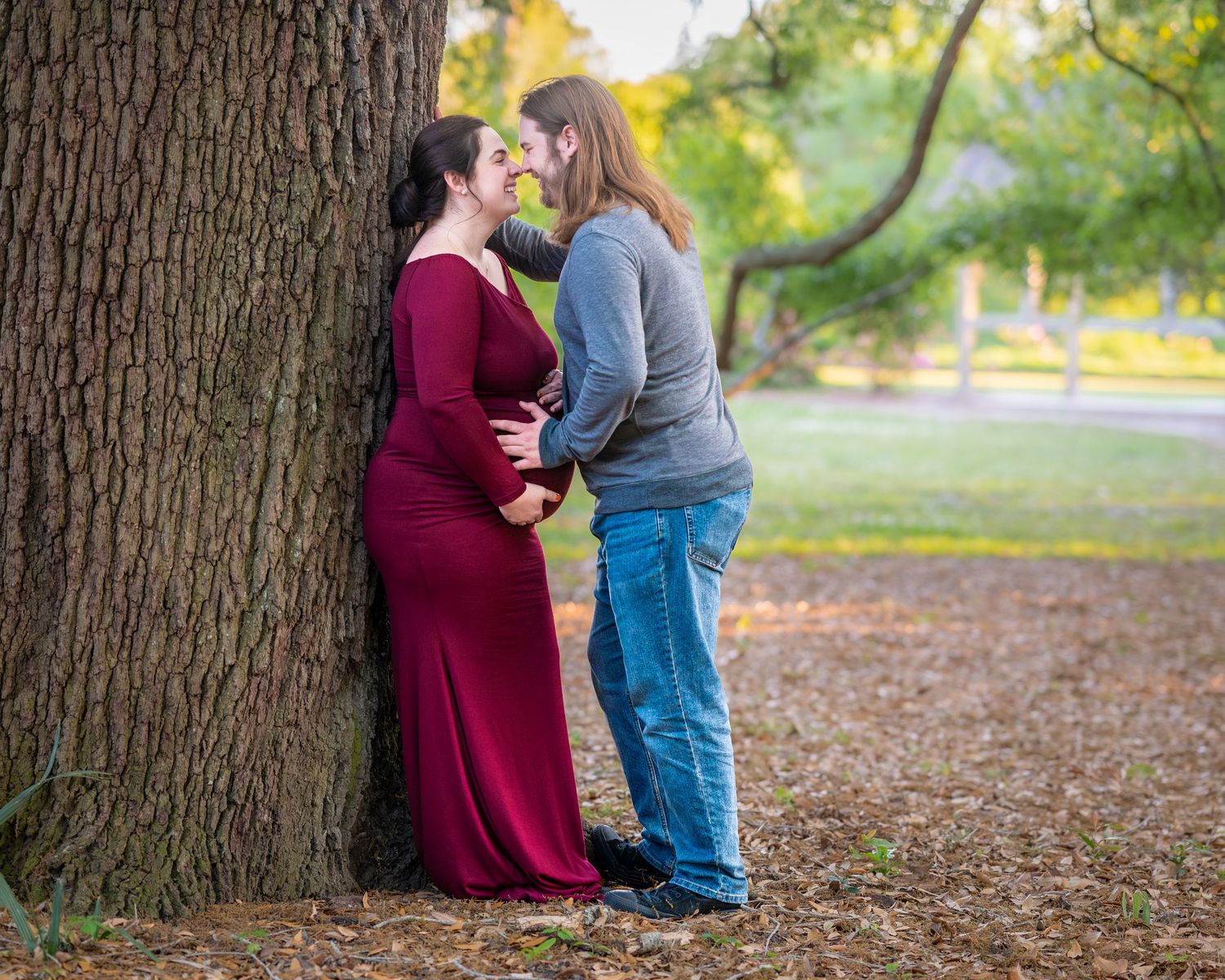 Lucy stands with her back against a large tree while her husband leans and kisses her during their maternity session at Hampton Park
