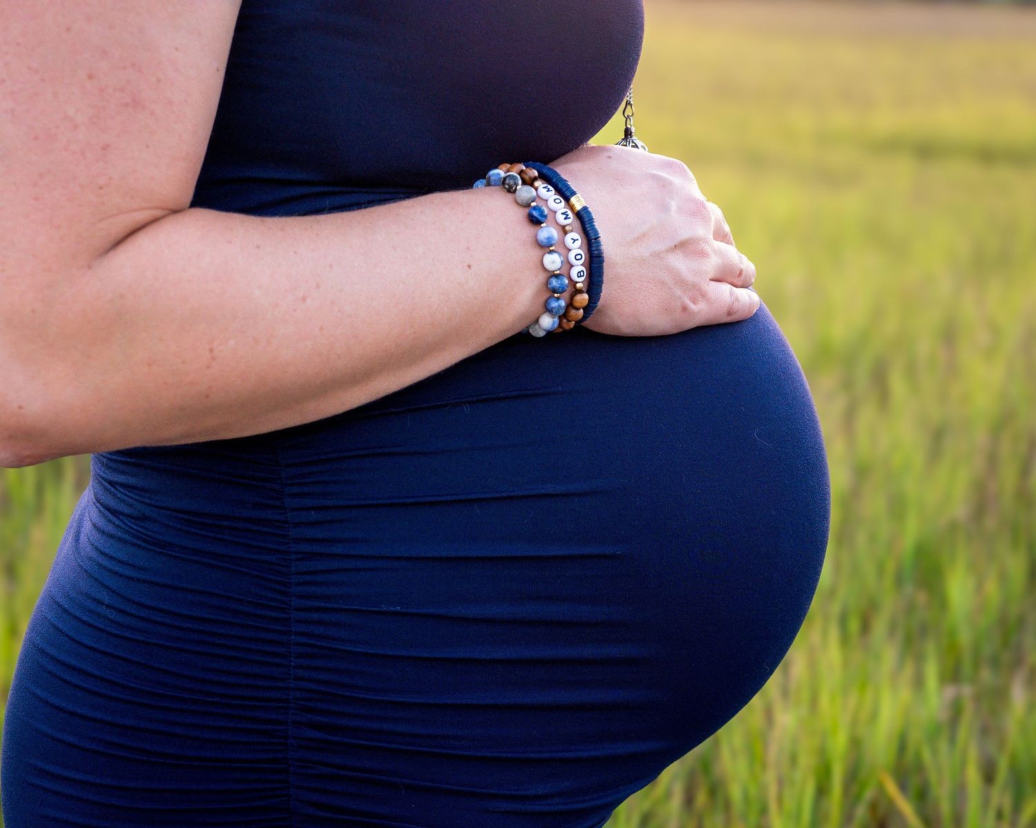 Close up of a pregnant belly with decorative bracelets against marsh grass background.