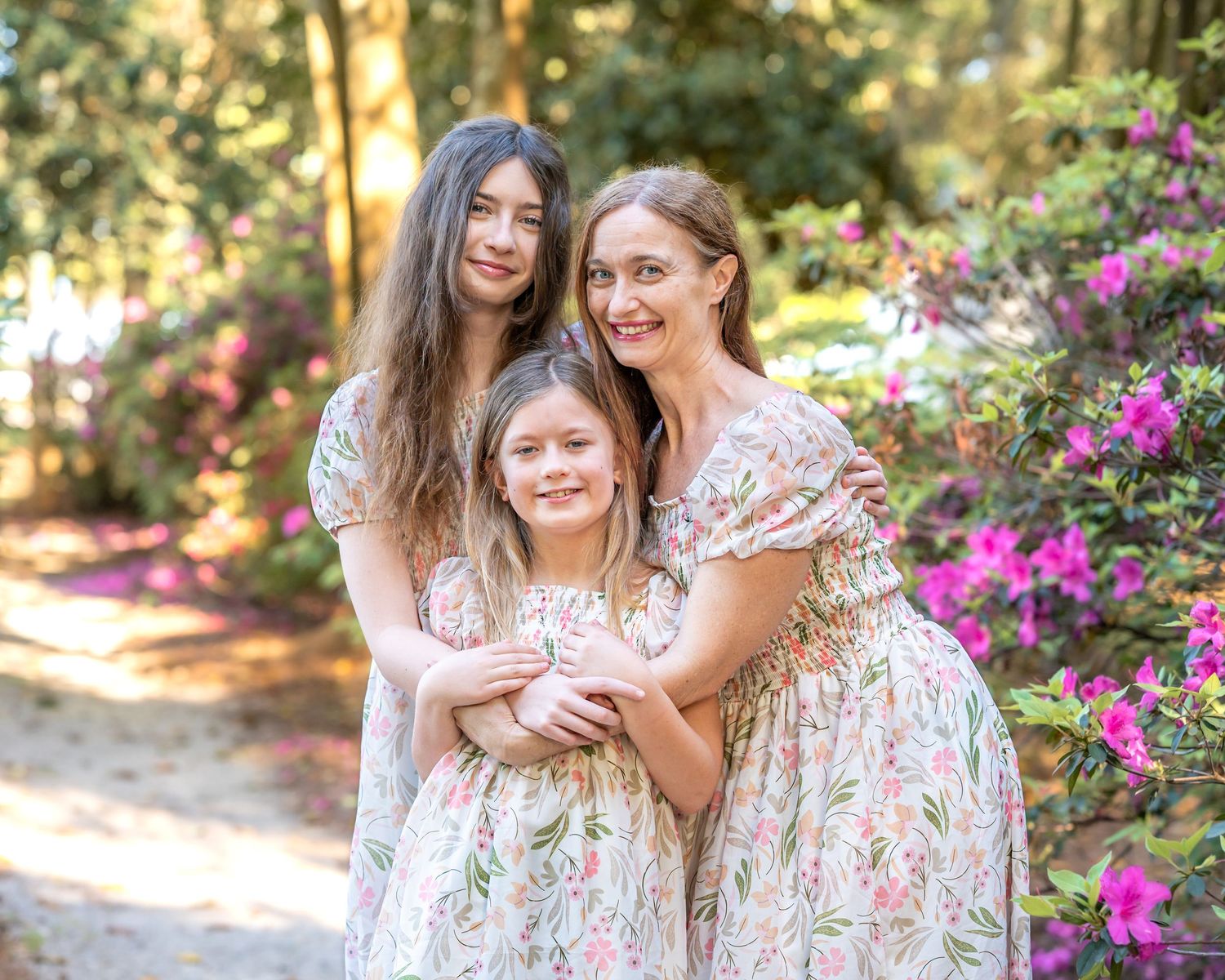 Three females in matching floral dresses pose together in a blooming garden with pink azaleas and tall trees.