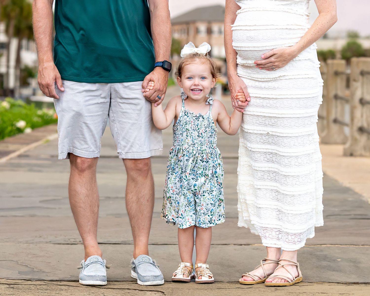 Young child in a floral summer dress stands between two adults wearing white on an outdoor walkway during their family maternity session