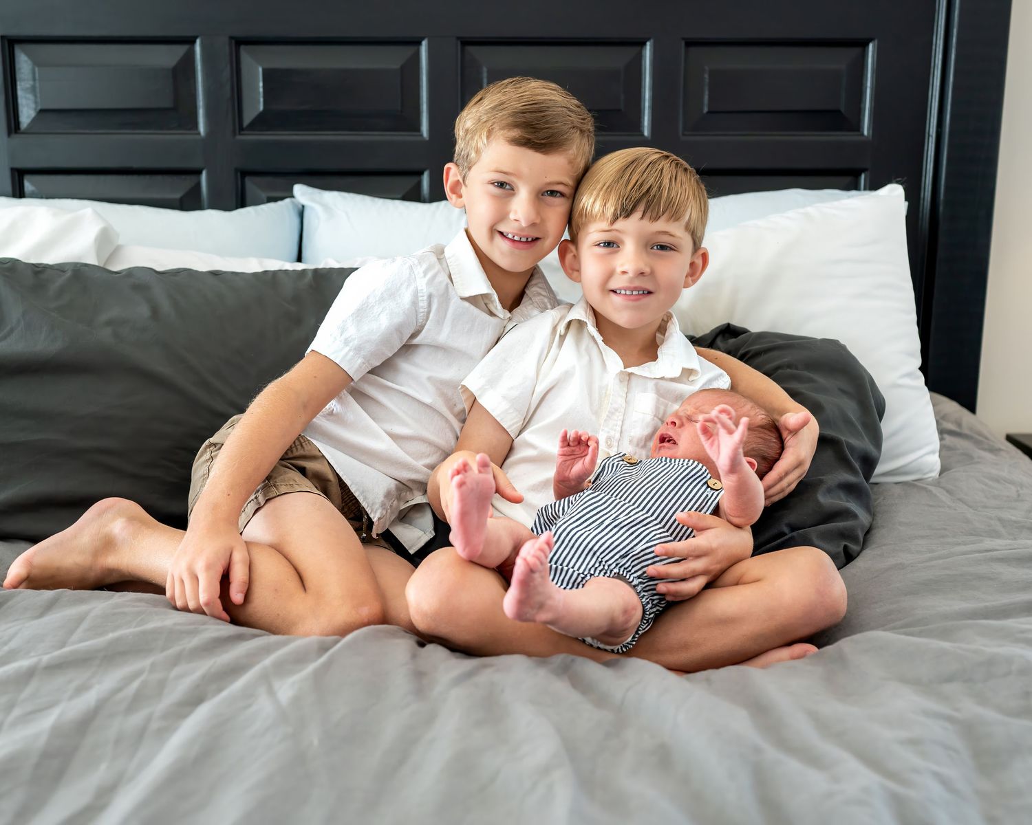 Siblings sit together on bed with newborn baby.