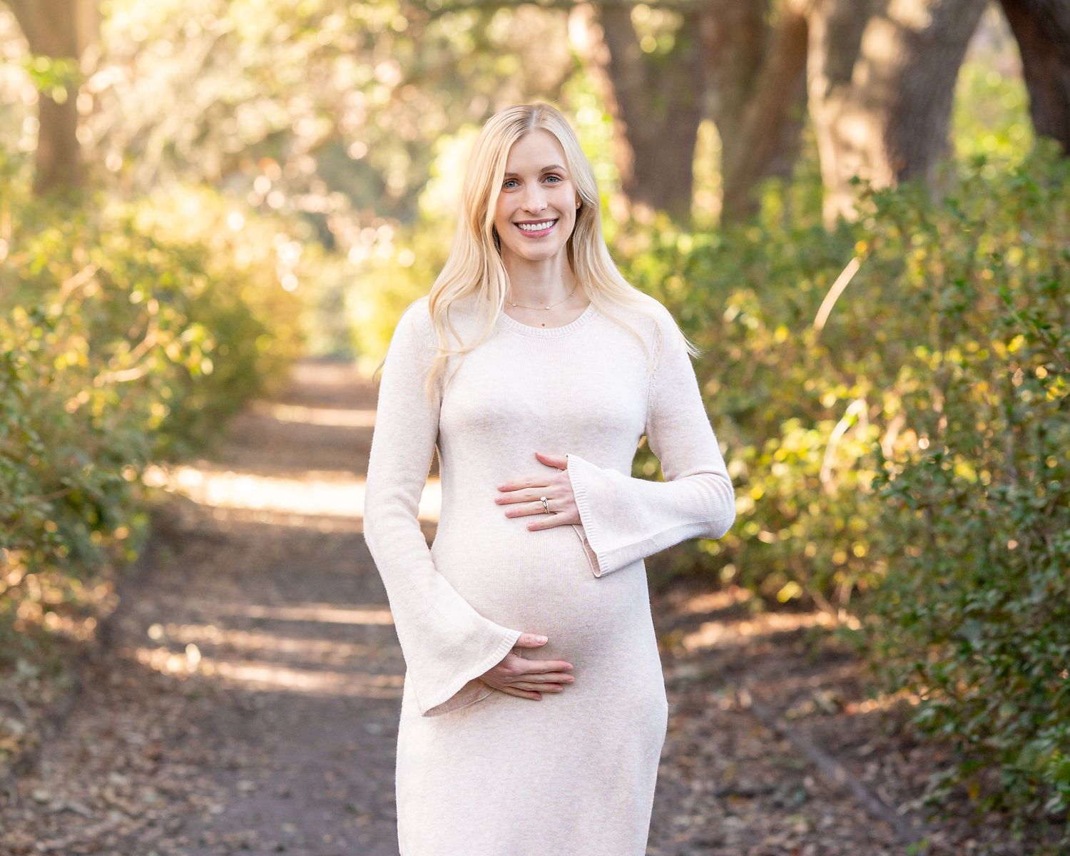 Maternity photo shoot in a sunlit wooded path wearing a flowing cream dress.
