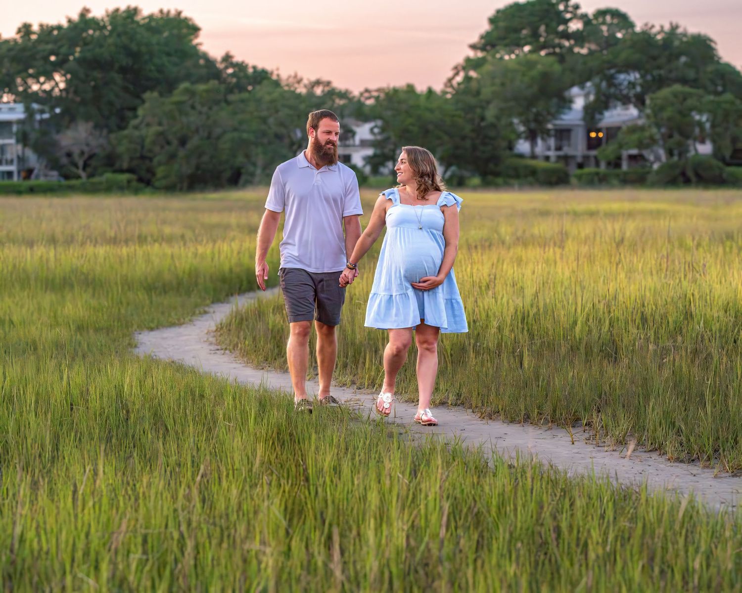A couple walks along a winding dirt path through golden marsh grass at sunset.