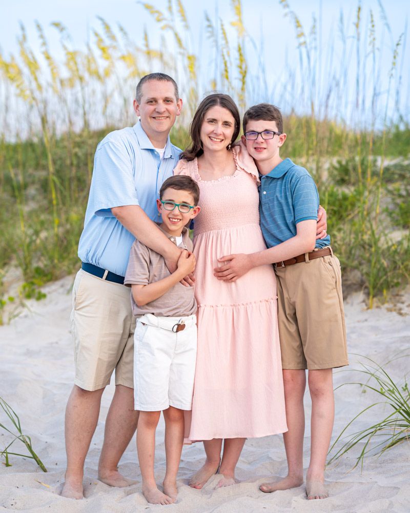 Family of four posing together on a sandy beach with sea oats in the background during a professional photo session.