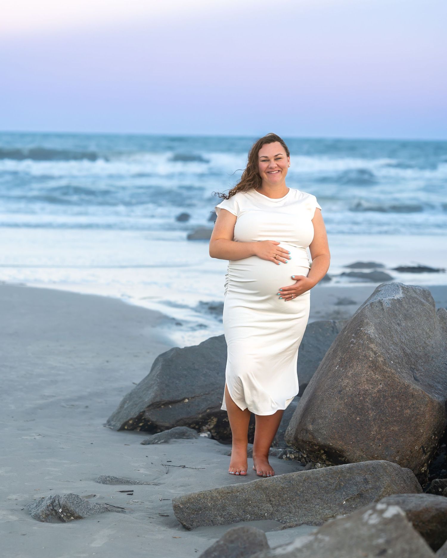 Pregnant woman in white dress smiling on a sandy beach at dusk, cradling her baby bump near large rocks.