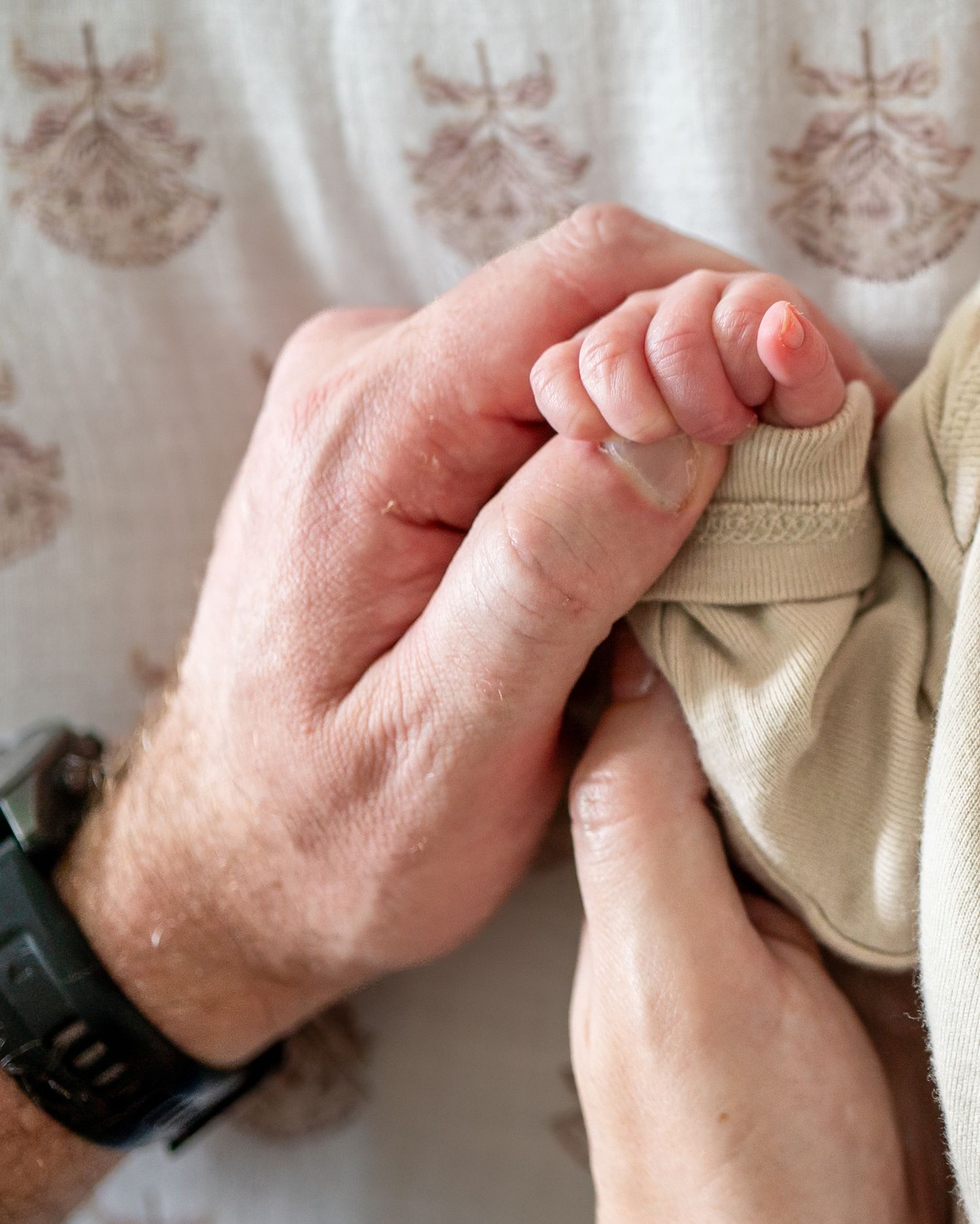 Adult hands gently holding a newborn baby's tiny fist, with a watch visible on the wrist.