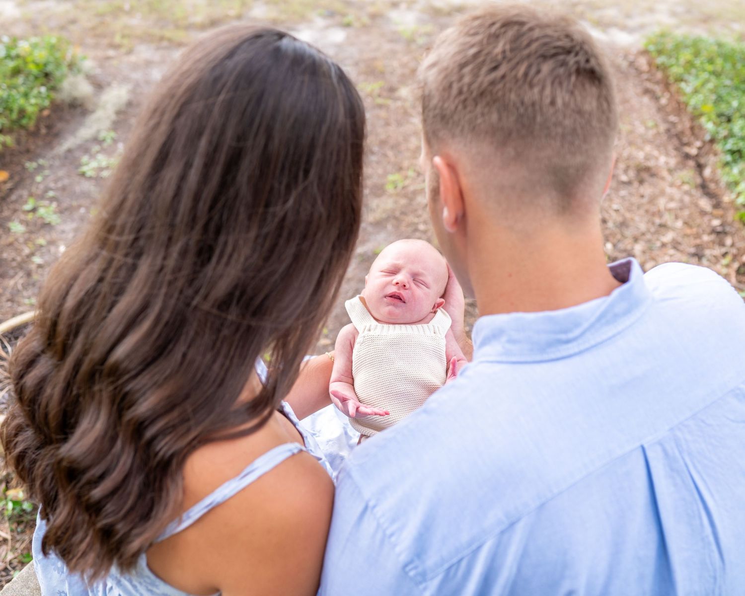 Picture taken from behind parents with a small opening the shows a newborn baby boy wearing a cream colored romper