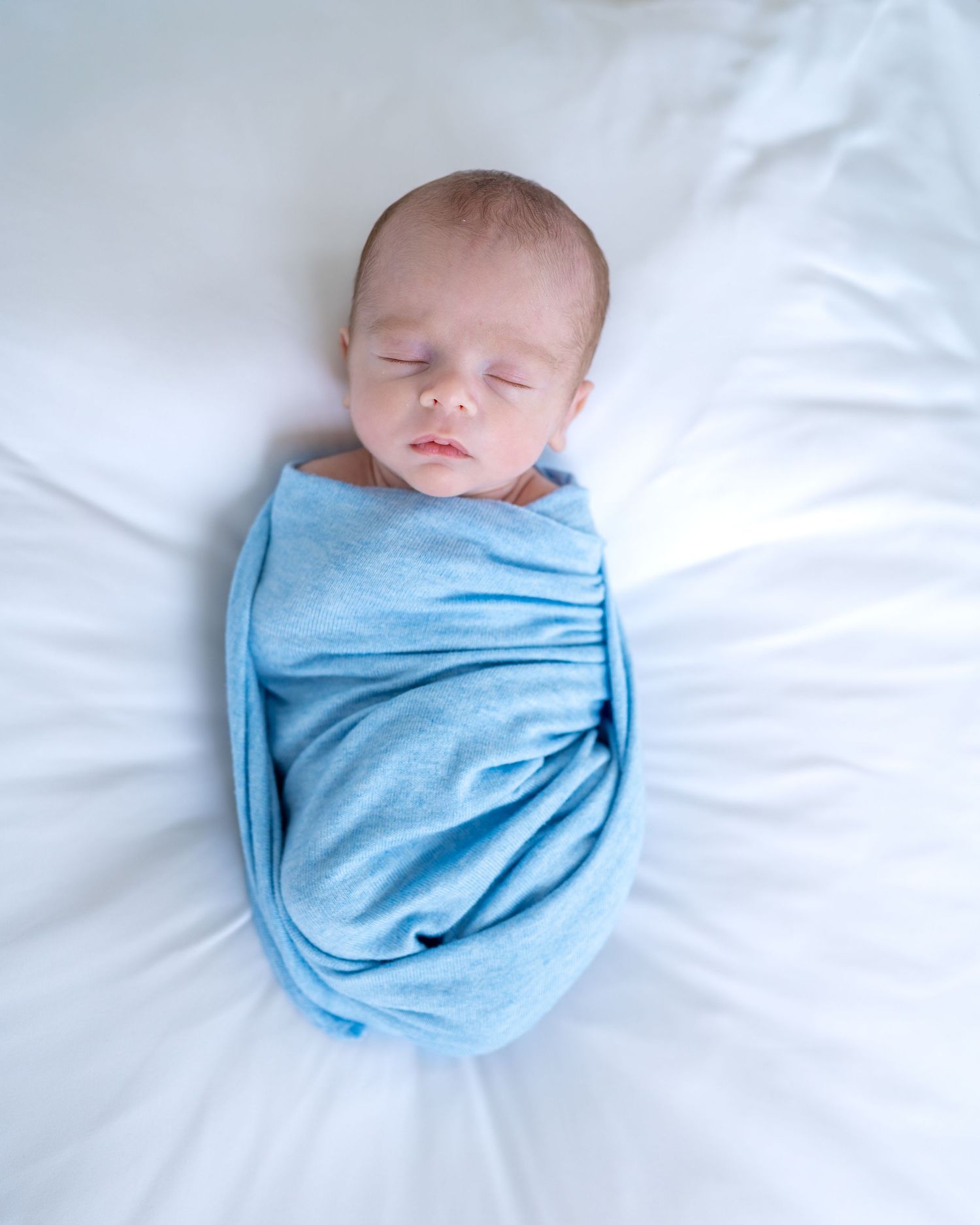 A peaceful newborn baby swaddled in a light blue wrap, sleeping on a white soft blanket.