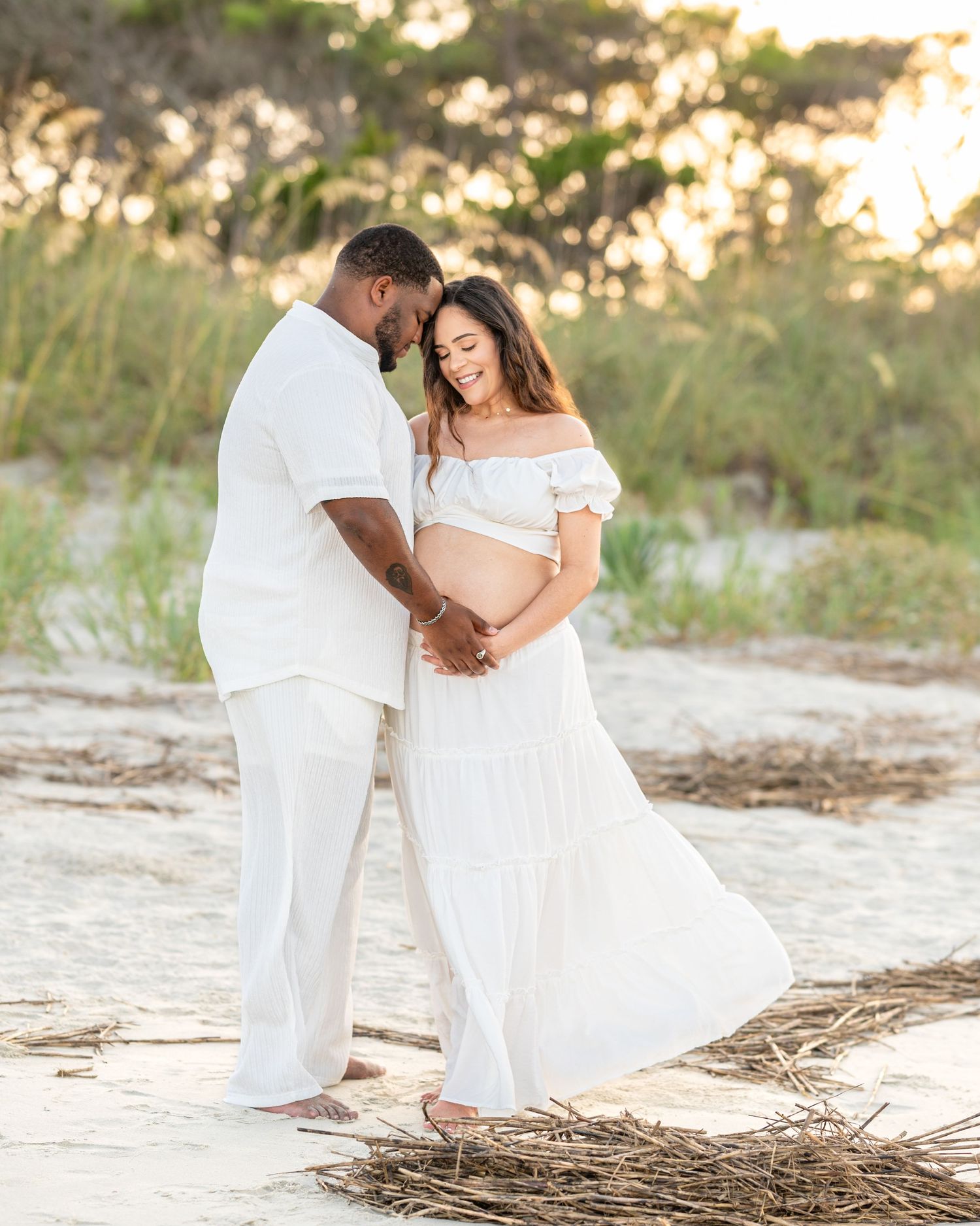 A couple in white outfits share an intimate moment on a beach during golden hour.