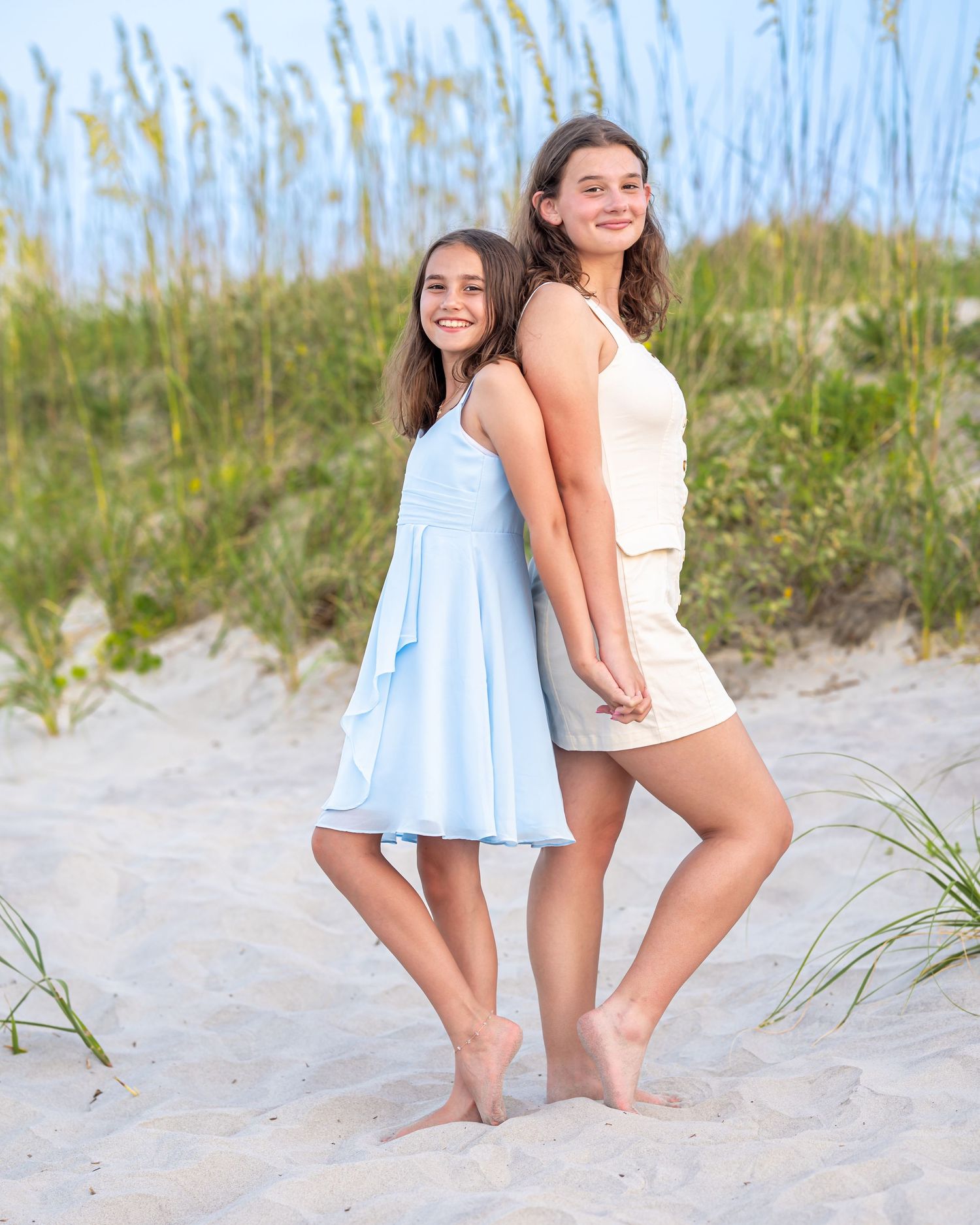 Two smiling girls in summer dresses pose back-to-back on a sandy beach with sea oats in the background.