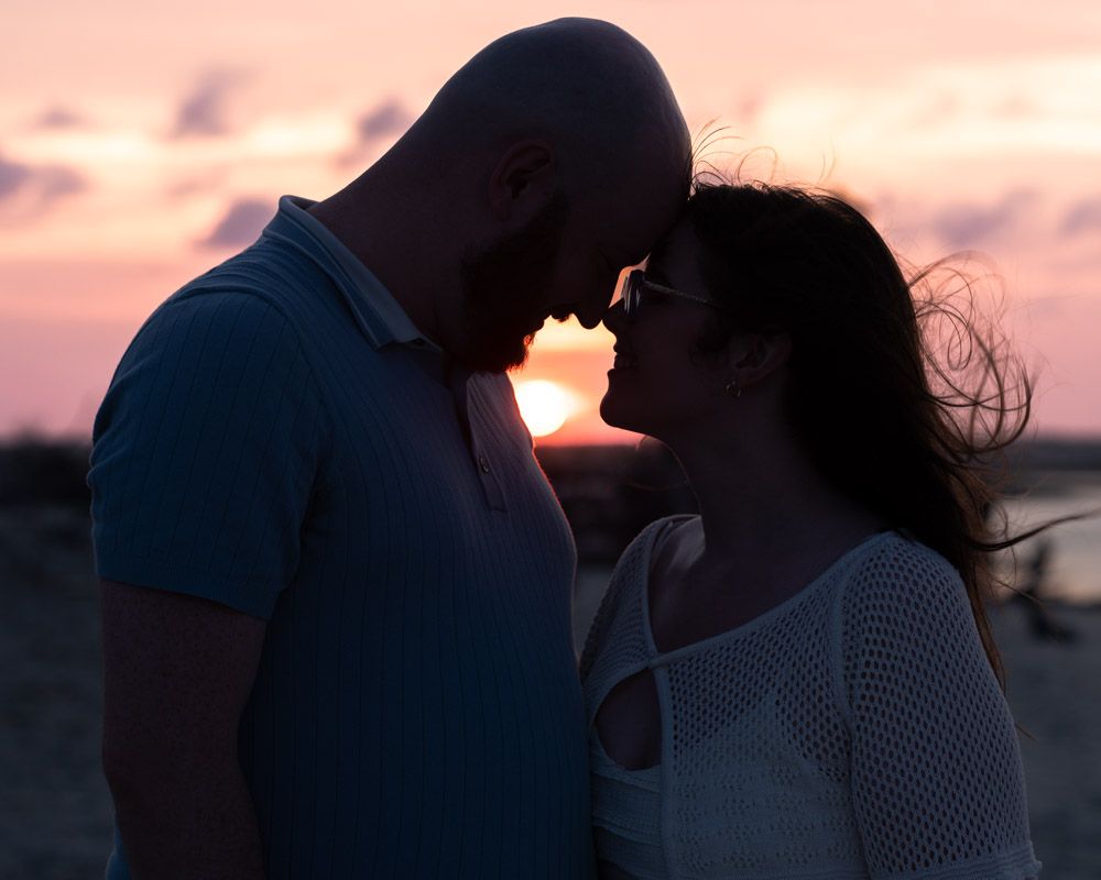 Romantic couple silhouetted against a vibrant pink and orange sunset sky, sharing an intimate moment on a beach.