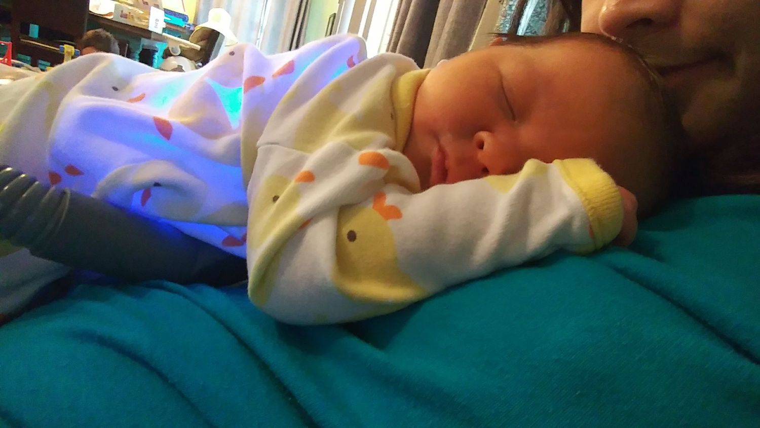 A newborn baby lies sleeping on his mom while wrapped in a biliblanket