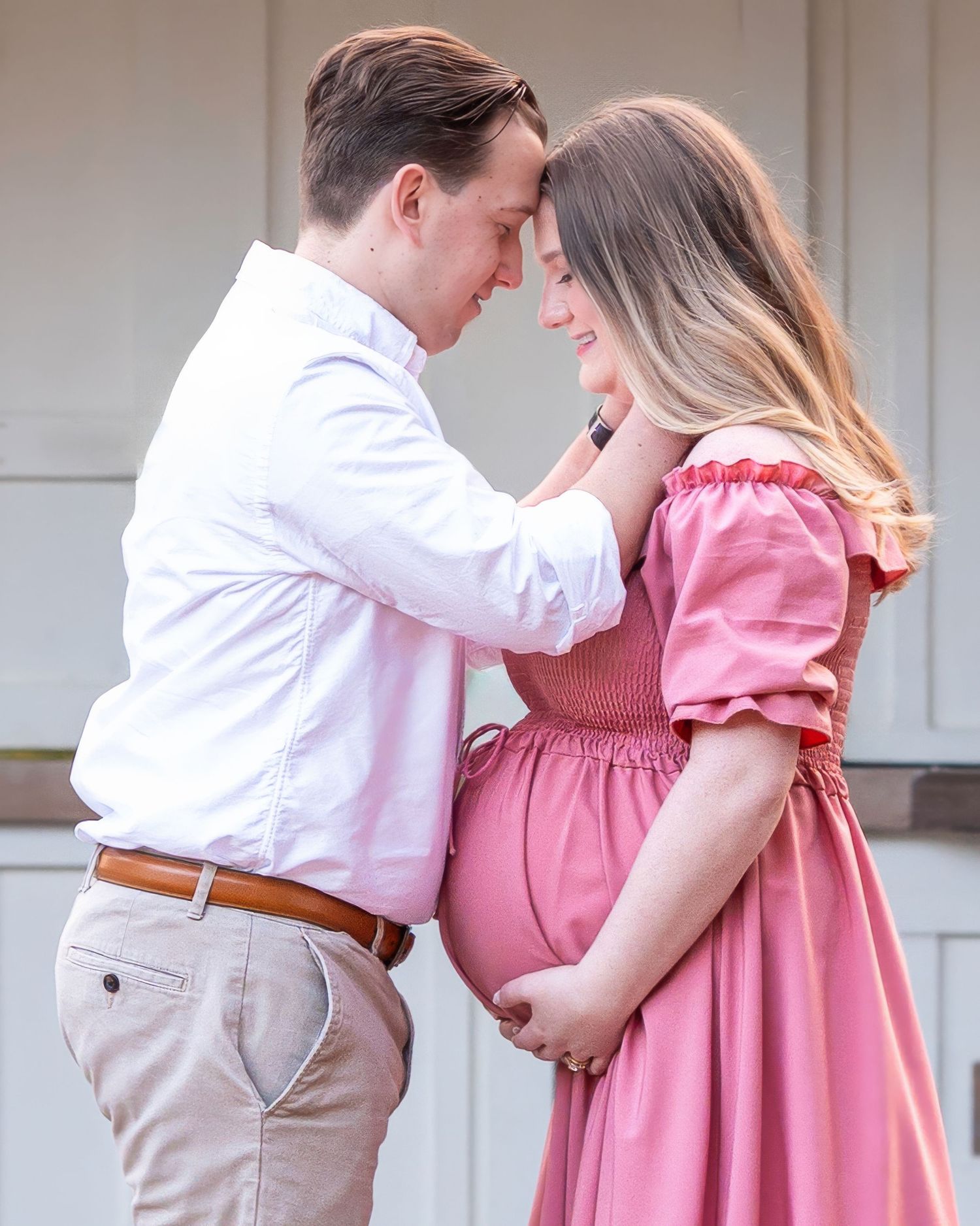 A couple in matching pink and white outfits embraces lovingly in a romantic outdoor maternity photoshoot.