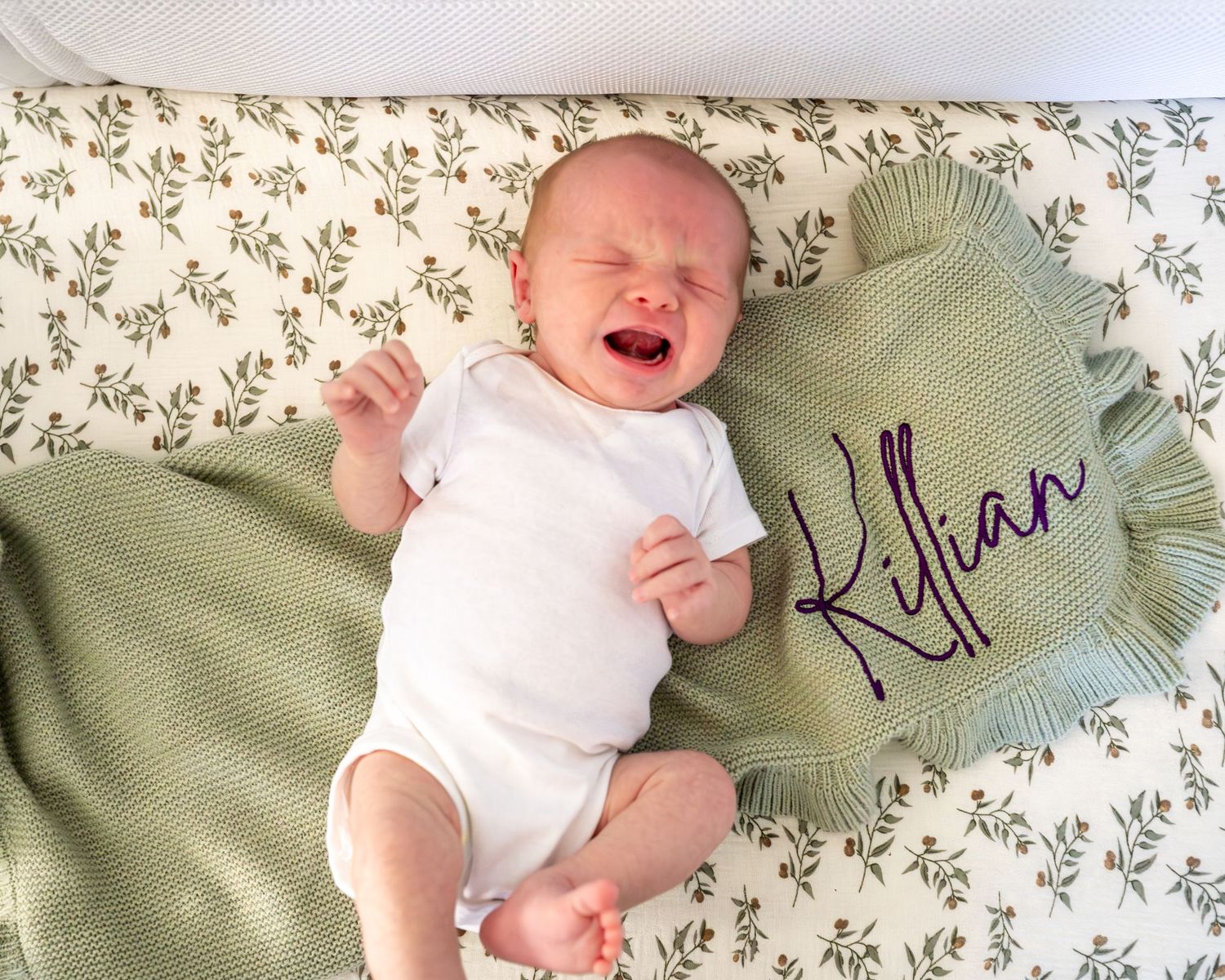 A smiling baby in white onesie lies on floral bedding next to a green pillow with embroidery.