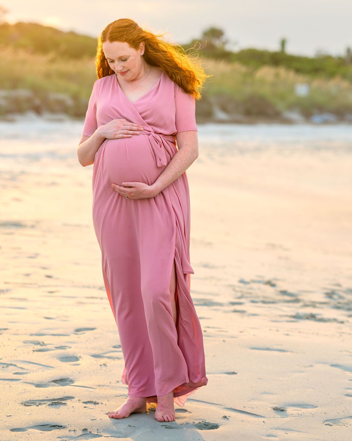 Expecting woman with beautiful red hair, wearing pink dress, looking down at her baby bump on the beach