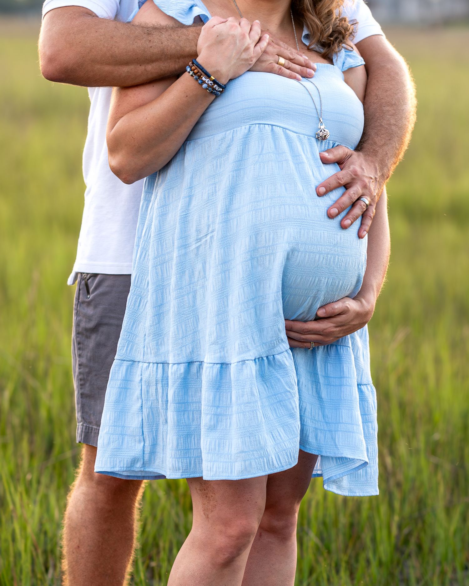 Close-up of a pregnant woman in a light blue dress being embraced from behind.