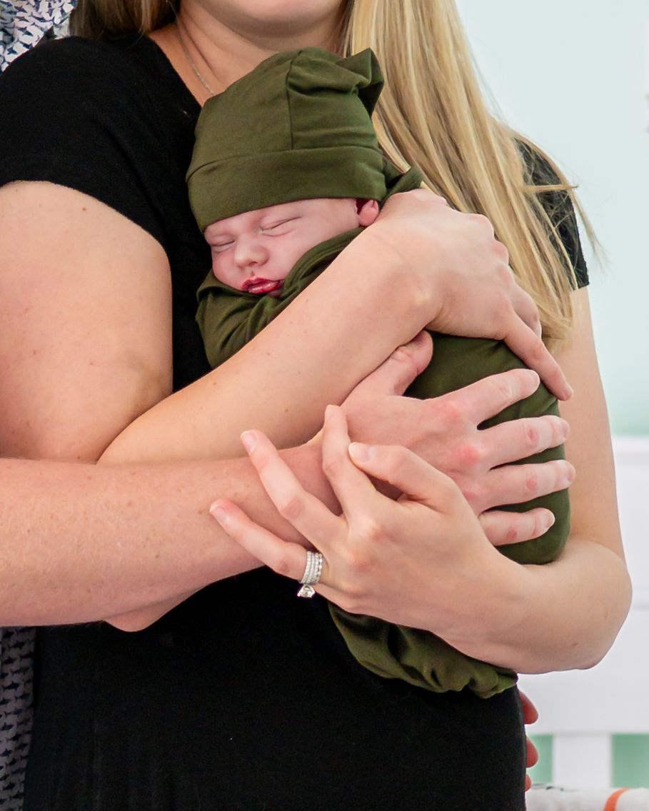 A woman in black holds a sleeping newborn wrapped in an olive green swaddle and matching knot hat.