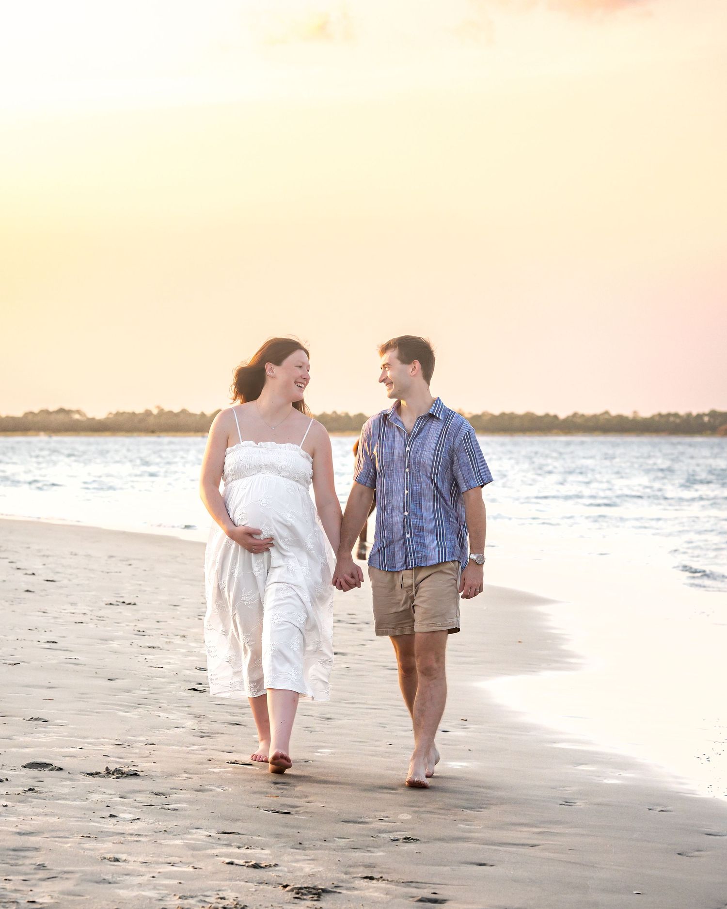 Pregnant couple walking hand-in-hand on a sandy beach at sunset, woman in white dress cradling baby bump.