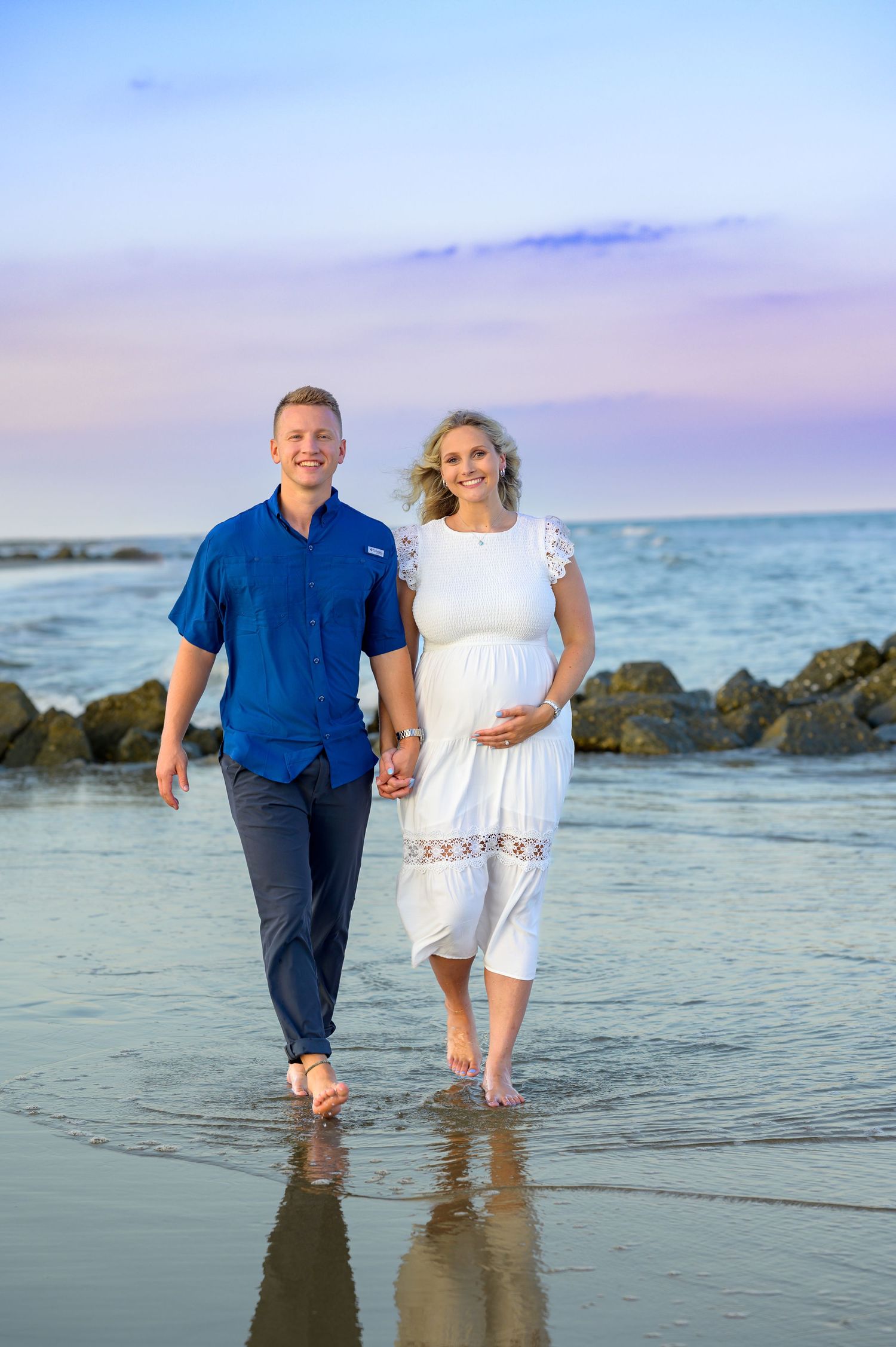Expecting couple holding hands walking towards photographer. blue and purple sunset skies behind them