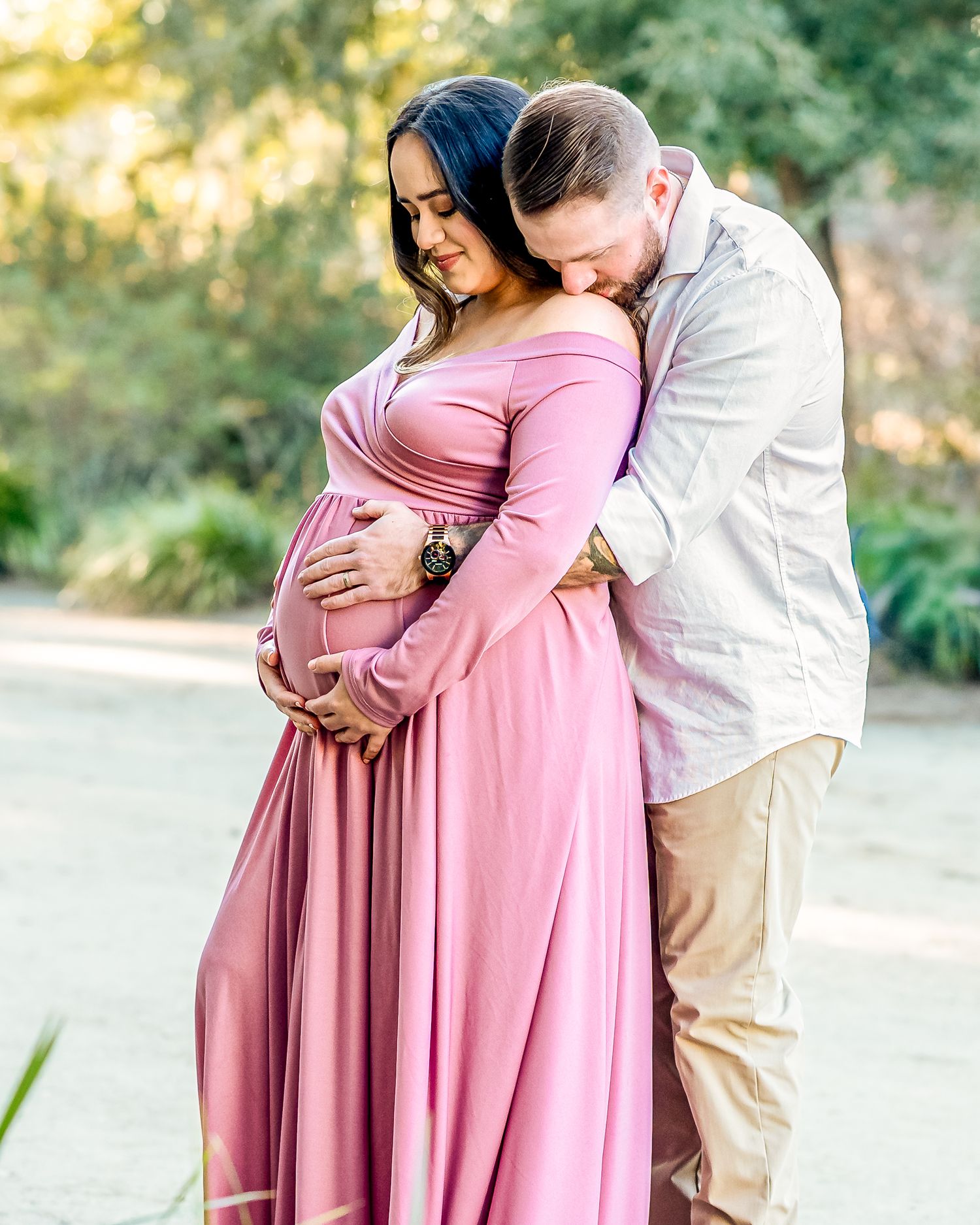 A couple shares an intimate embrace outdoors, with the person wearing a flowing pink dress during a maternity photo session.