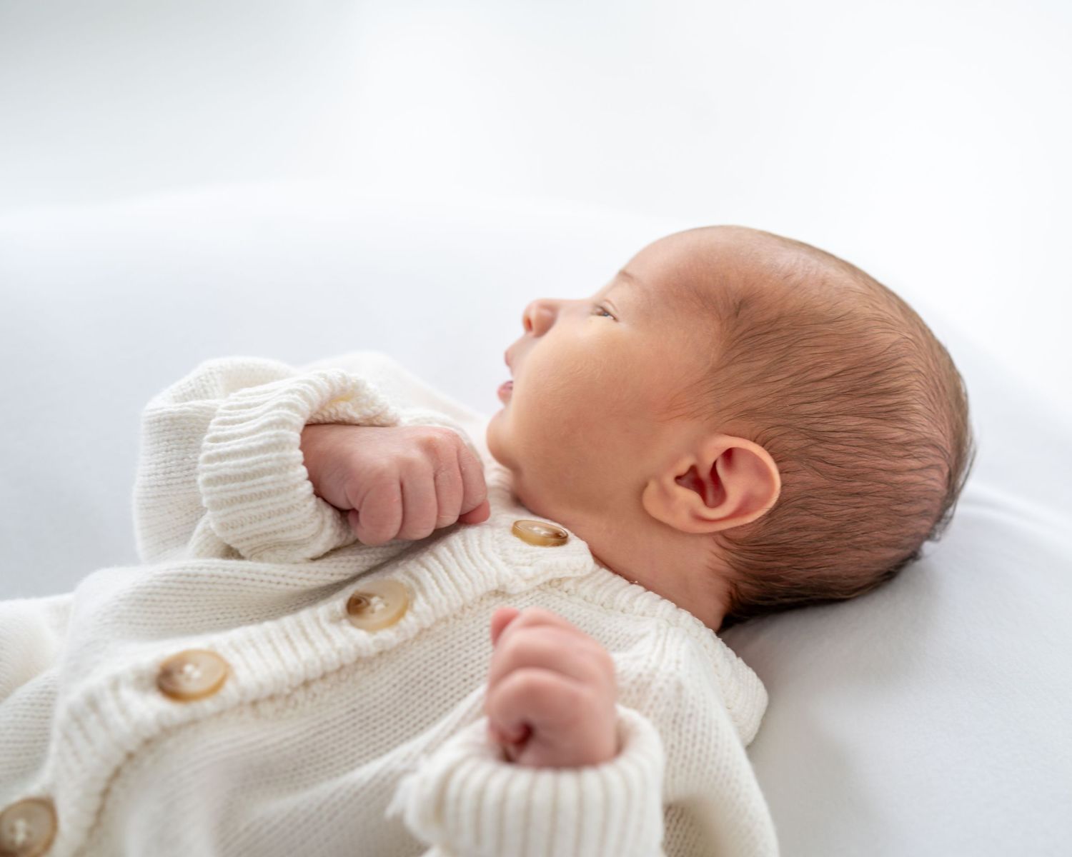 A sleeping newborn baby wearing a white knit sweater lies peacefully on a white blanket.