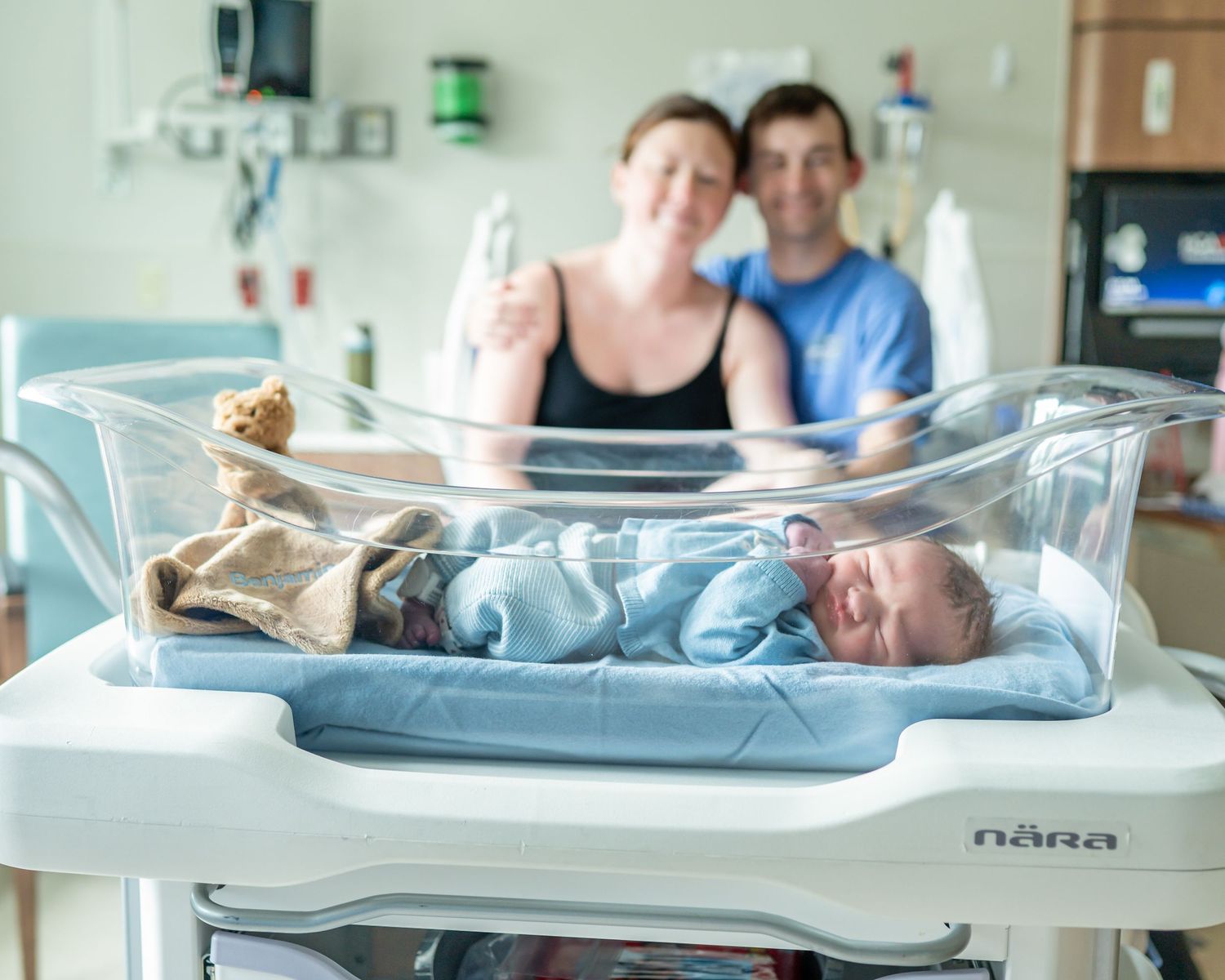 Newborn baby resting in a hospital bassinet with proud parents smiling in the background.