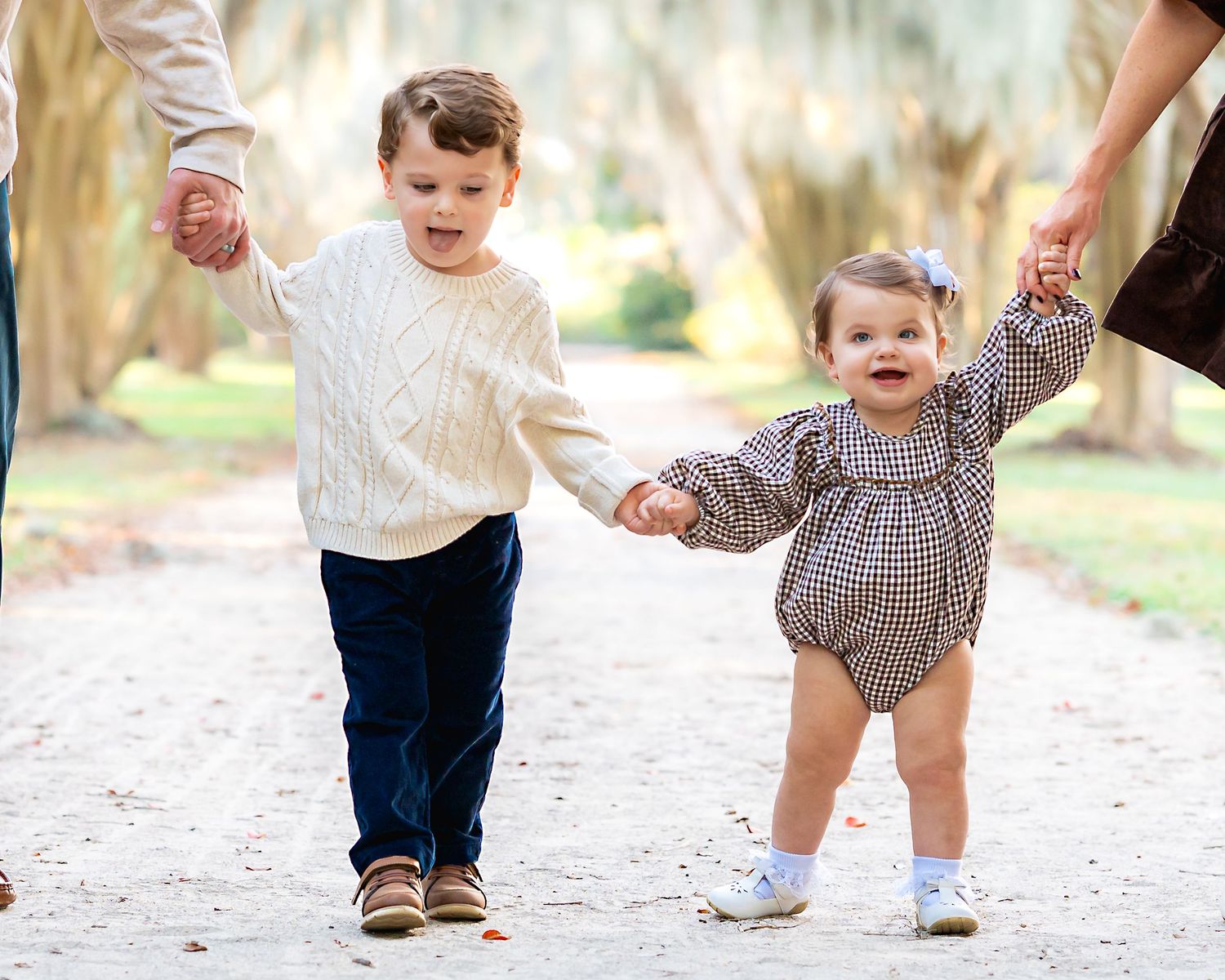 Two young siblings holding hands with parents on a tree-lined path, dressed in coordinating fall outfits.