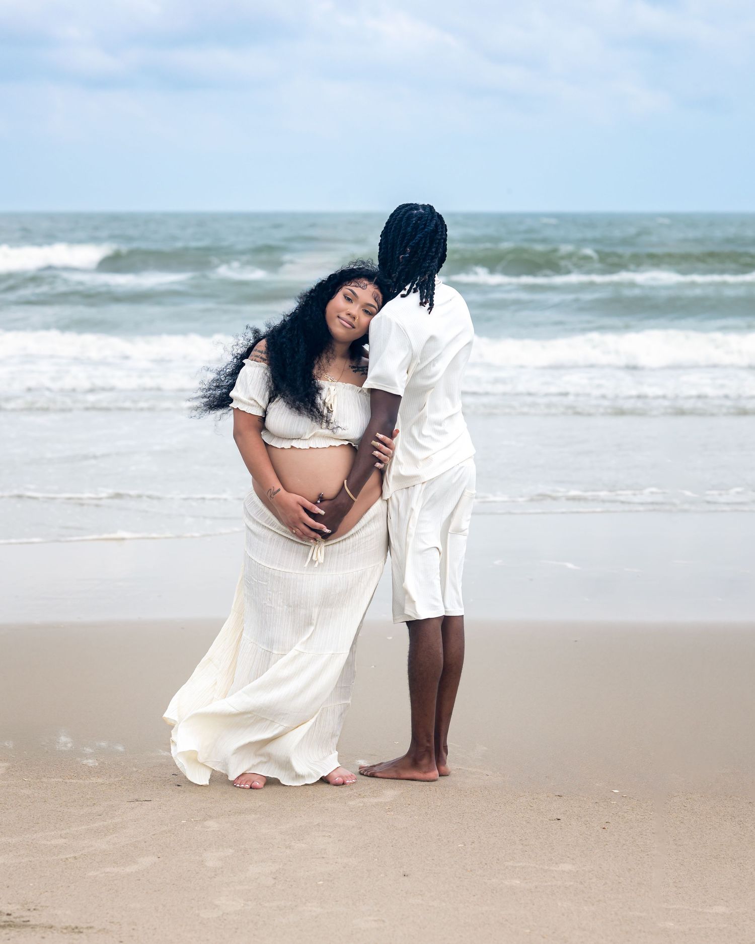 Pregnant couple in white outfits embracing on a beach, waves in the background, maternity photoshoot.