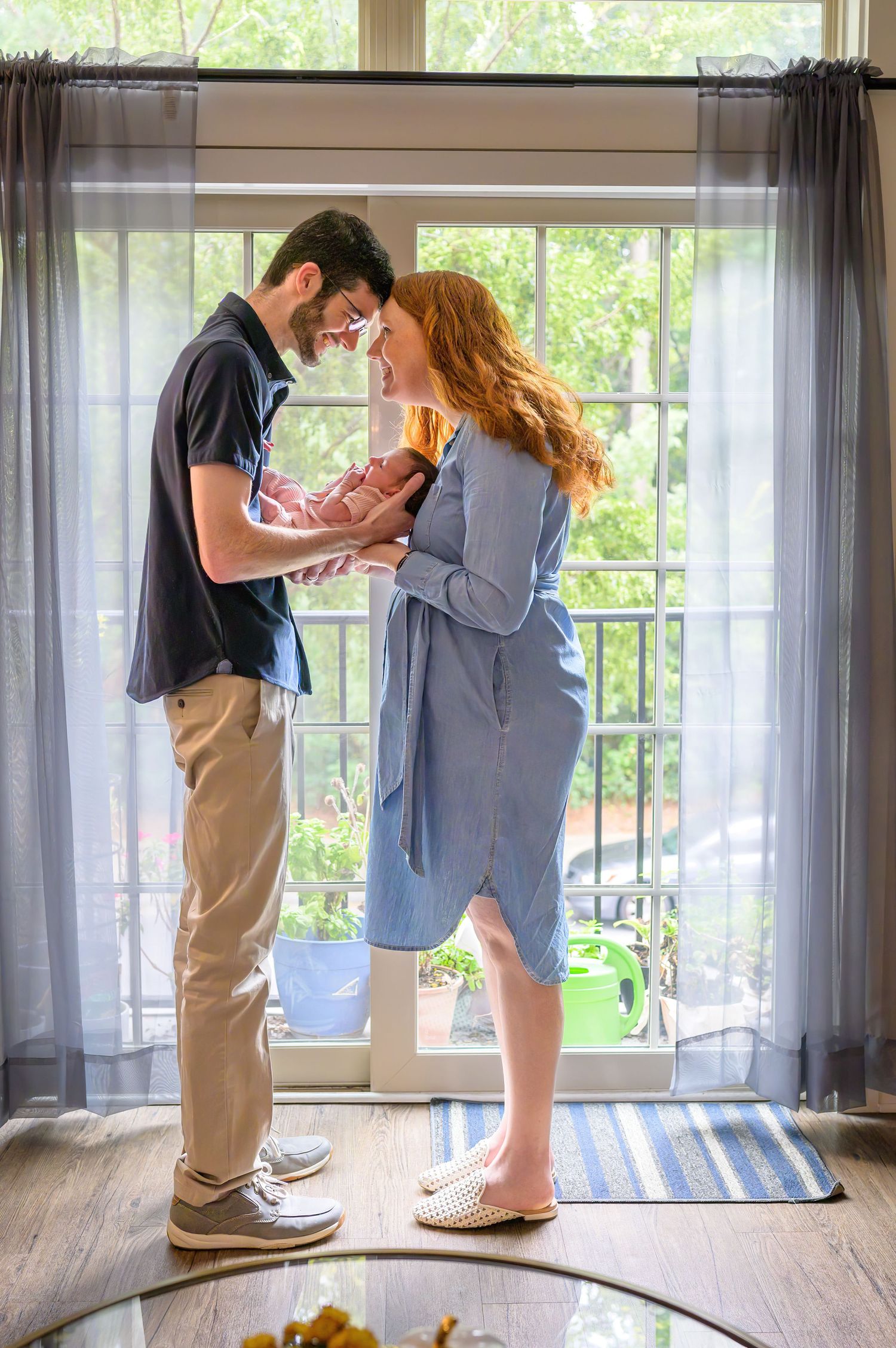New parents share a tender moment holding their newborn baby by a sunlit window in their home.