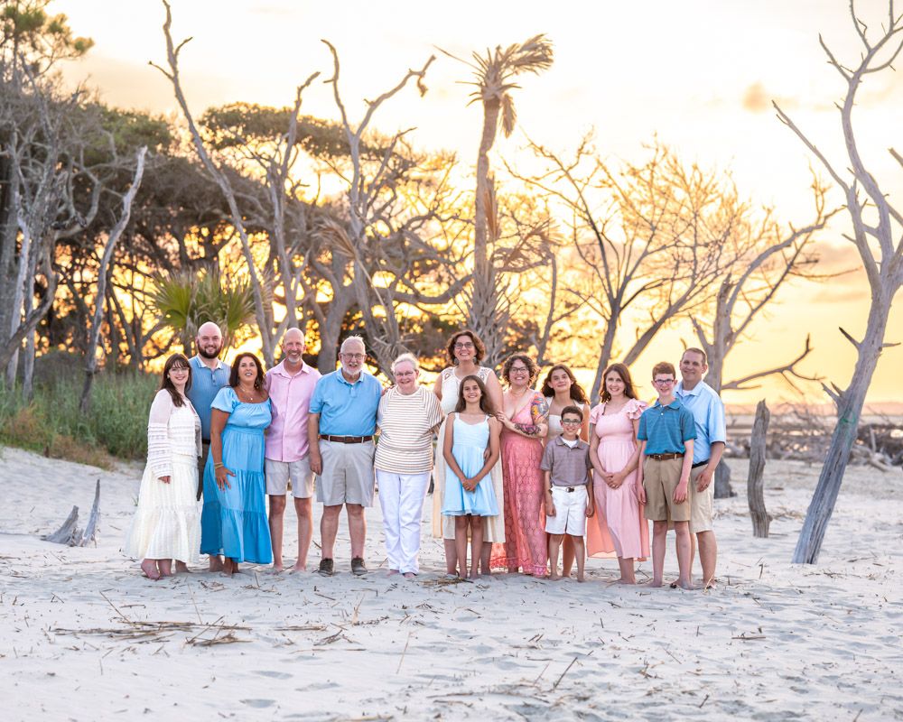 Large family group posing together on a sandy beach at golden sunset with bare trees and palm trees in background.