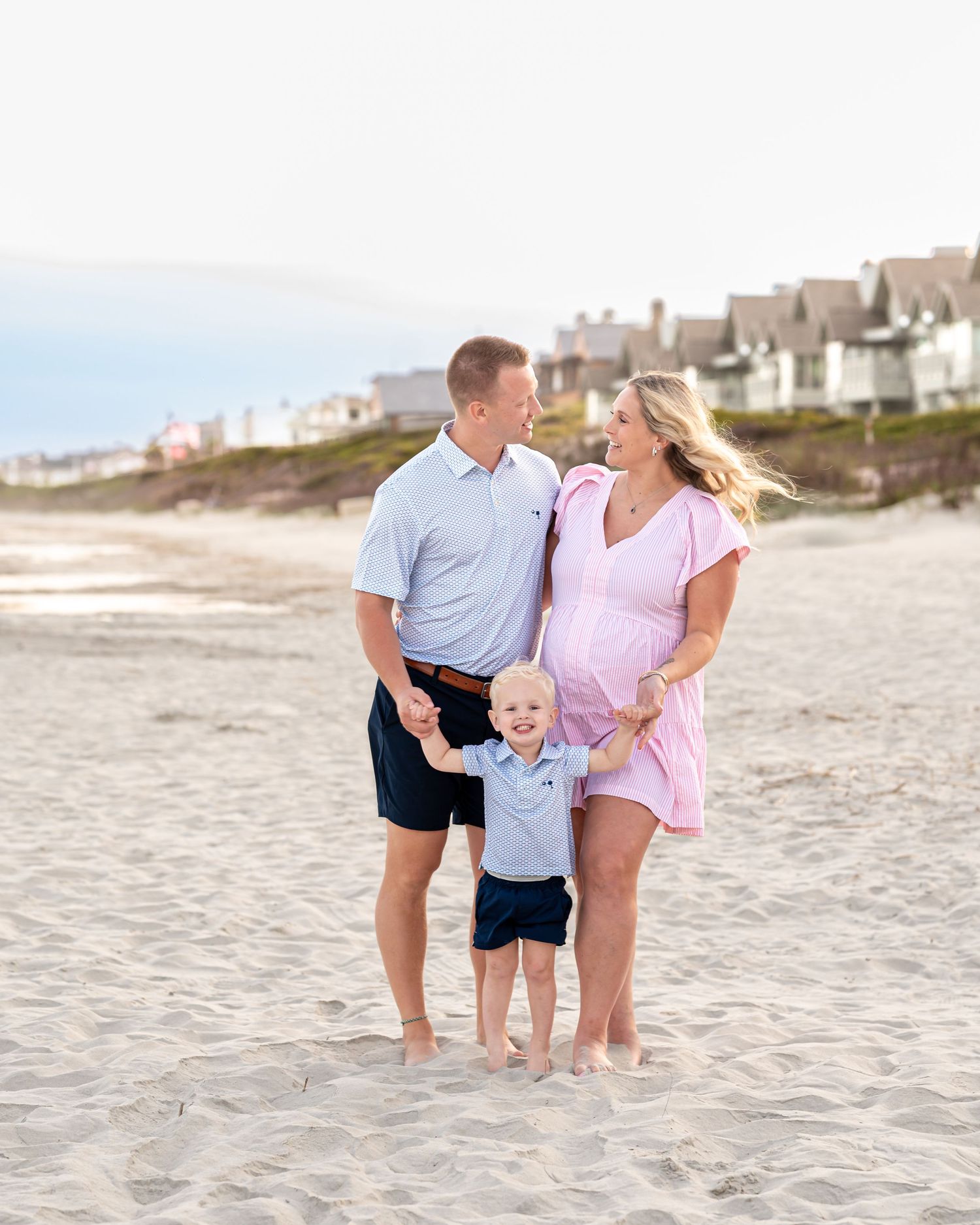 A happy family of three poses on a sandy beach, with a pregnant mother in a pink dress and a young toddler boy.