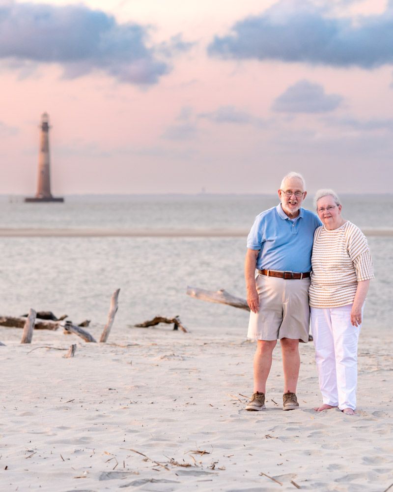 Elderly couple smiling on sandy beach at sunset with a lighthouse visible in the background.