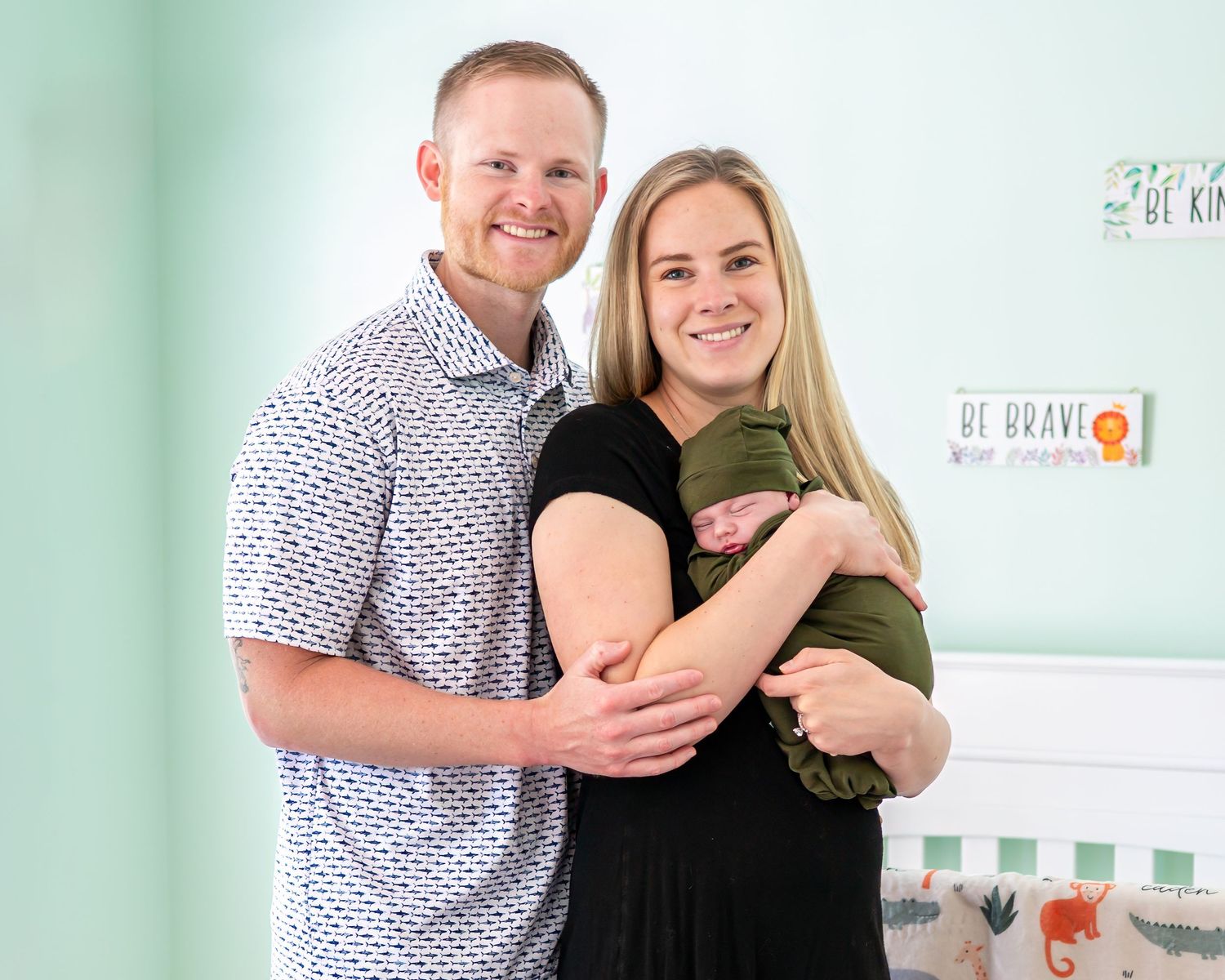 Happy couple smiling in nursery, holding newborn baby wrapped in green swaddle blanket.