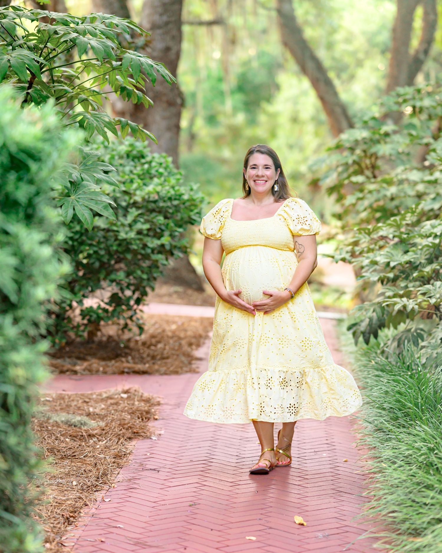 Pregnant woman in yellow eyelet dress cradling baby bump on brick garden path surrounded by lush green trees.