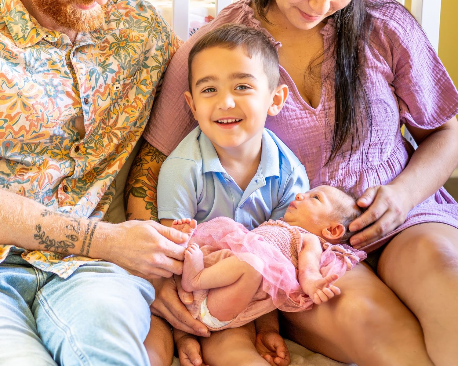Young child in blue shirt sits closely with adults while looking at newborn baby wrapped in pink blanket.