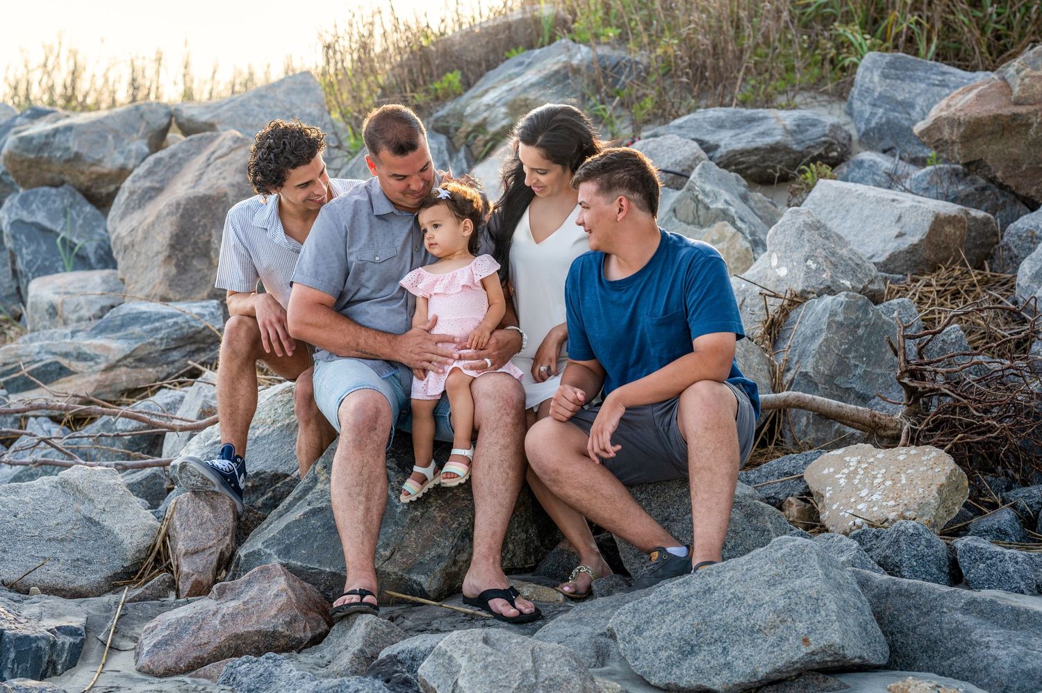 Large family including mom, dad, two teen sons, and todder little girl sitting together on beach rocks