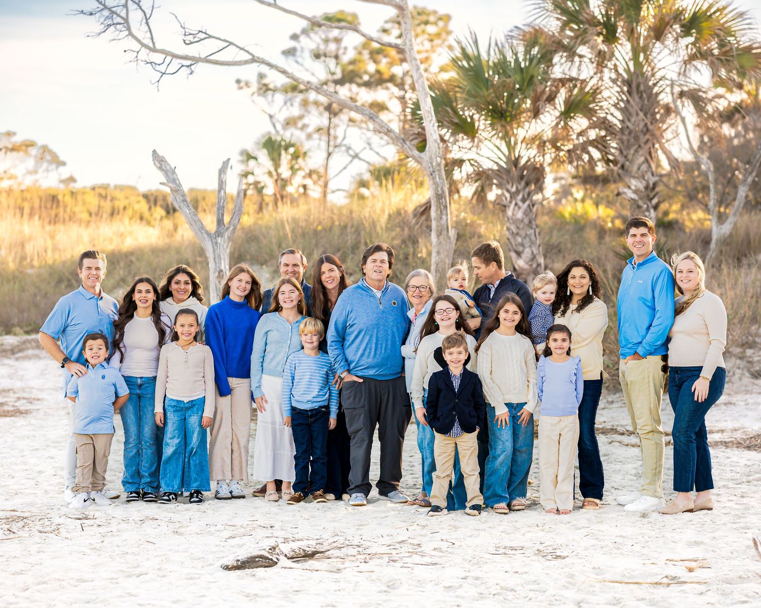A large extended family group poses together on a sandy beach at sunset wearing coordinated blue and white outfits.