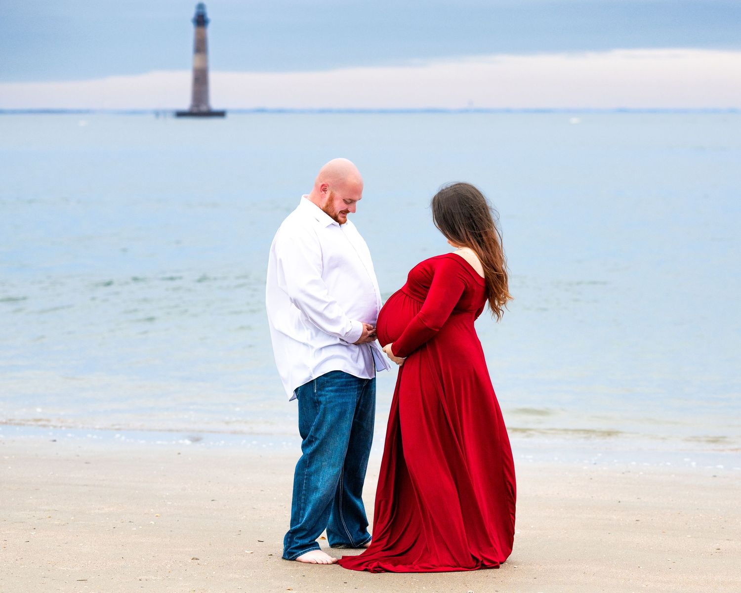 Two people in romantic pose on beach with lighthouse in background across calm blue waters on cloudy day.